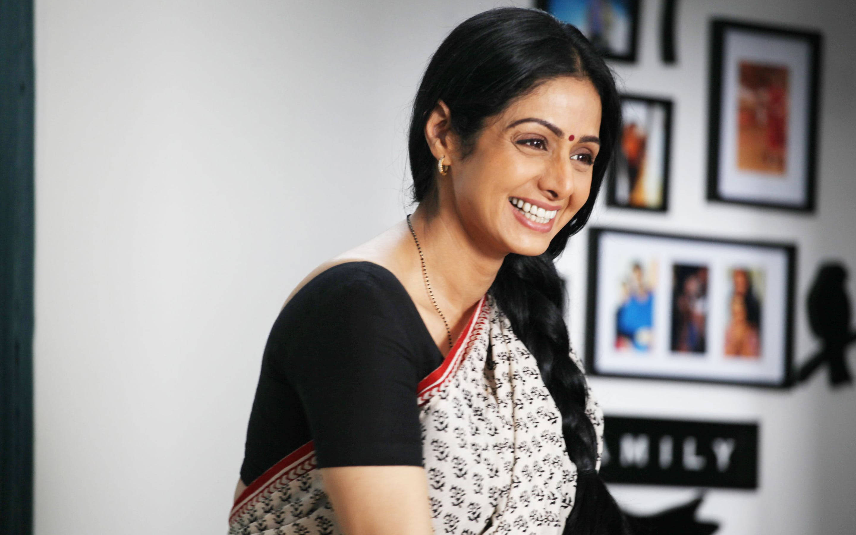 Sridevi 桌布 壁紙 — 一位微笑的女子身穿圖案莎麗，辮子與眉心點，站在一個有框照片和家庭標誌的房間裡。