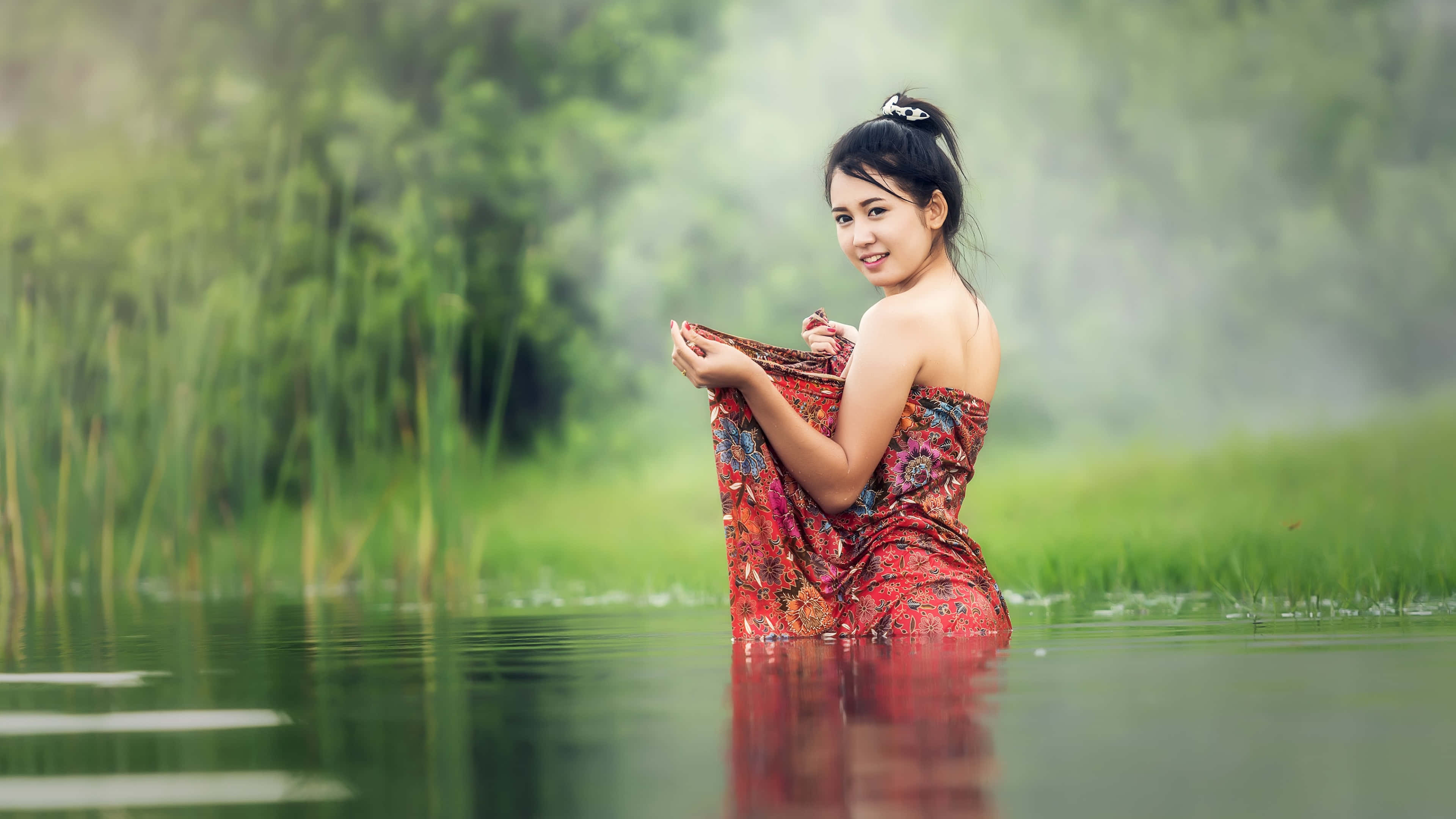 Teen Girl 桌布 壁紙 — 一位微笑的女子身穿彩色圖案連衣裙，站在平靜的水中，水深及腰，周圍是茂密的綠色蘆葦和霧氣瀰漫的樹葉。