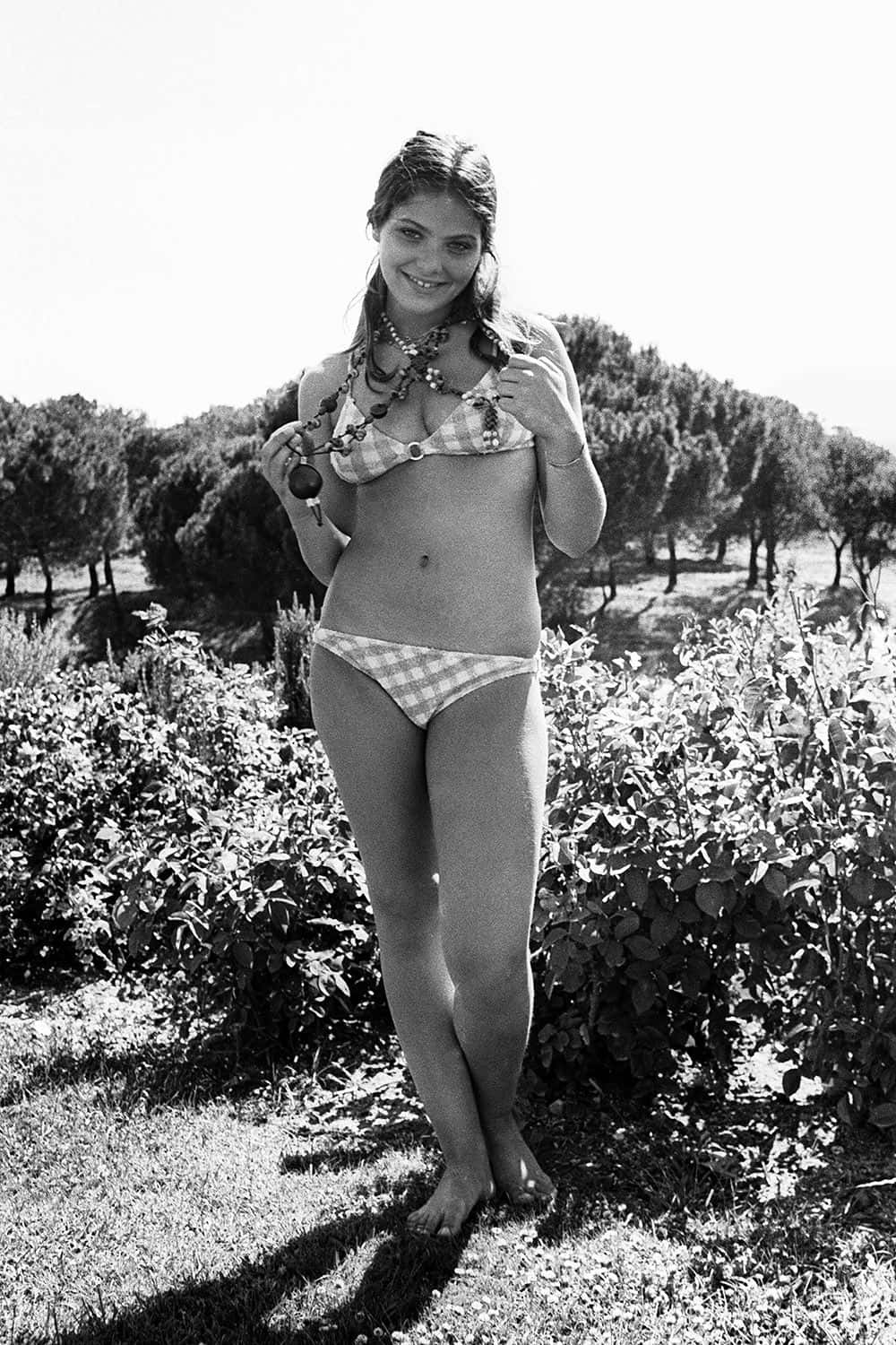 bikini model 桌布 壁紙 — 一位微笑的女子身穿格紋比基尼，赤腳站在陽光普照的花園中，戴著珠串項鍊。