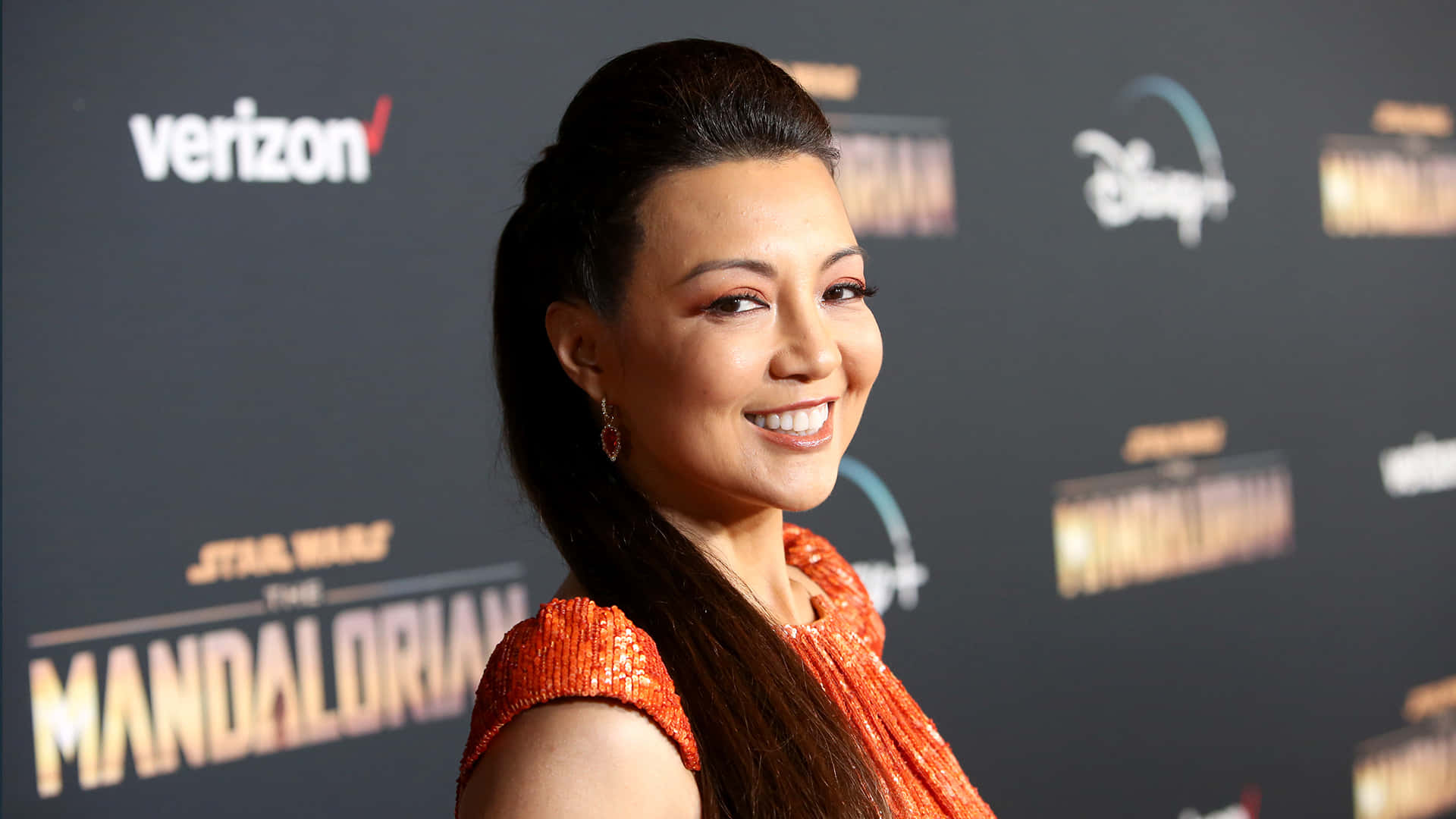Ming Na Wen 桌布 壁紙 — 一位微笑的女子身穿橙色亮片連衣裙，在《星際大戰：曼達洛人》活動中，於品牌背景前擺姿勢。