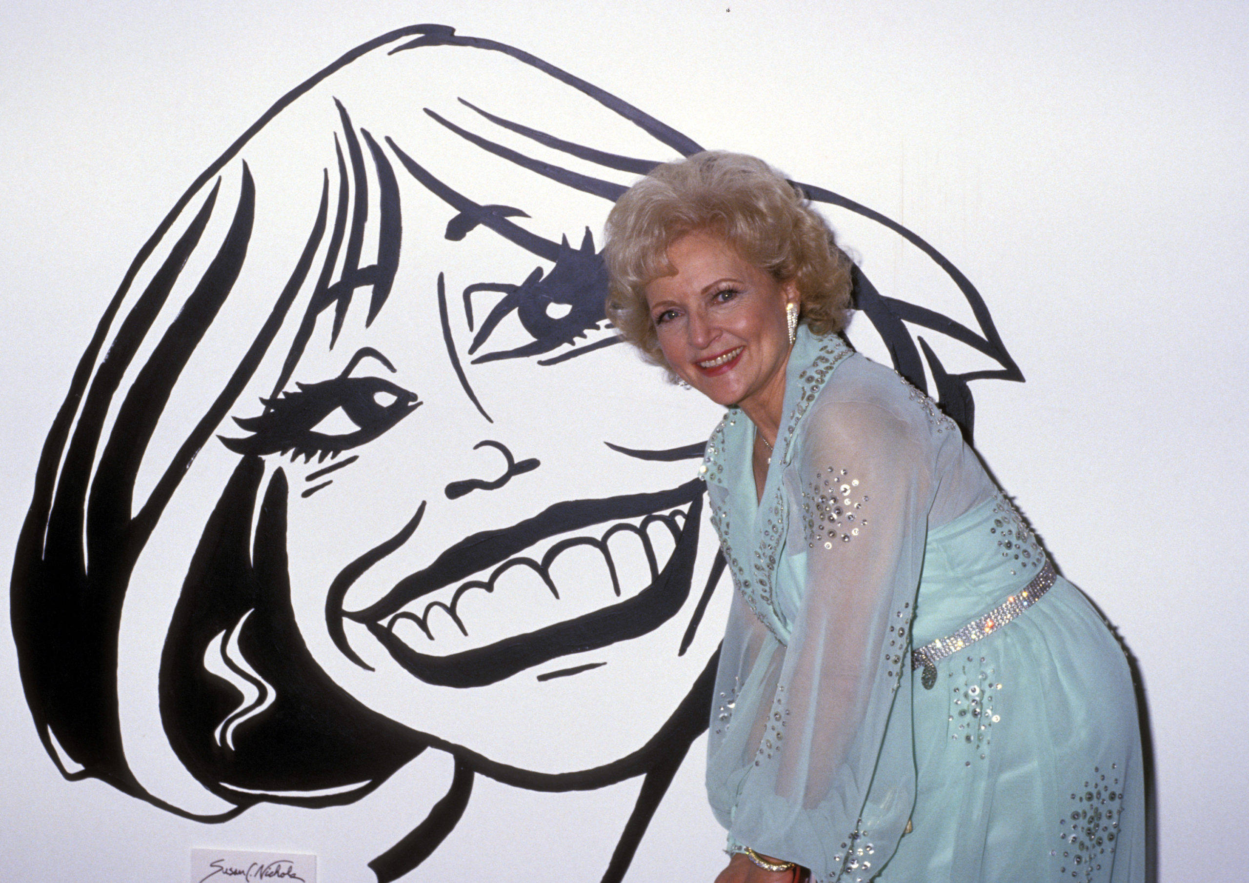 Betty White 桌布 壁紙 — 一位微笑的女子身穿淺藍色亮片連衣裙，站在一個她臉部的大型黑白卡通肖像旁邊擺姿勢。