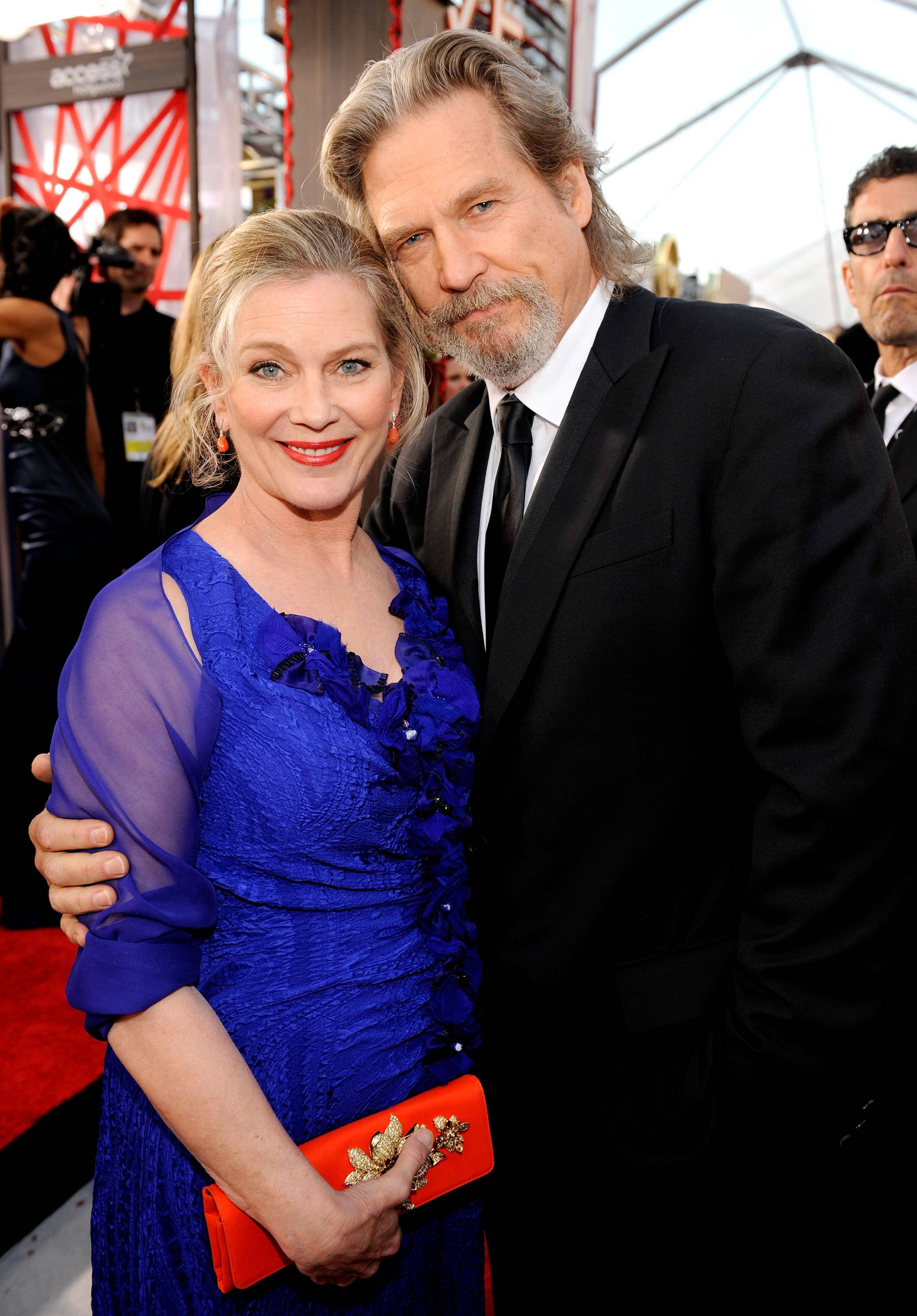 actor 桌布 壁紙 — 一位微笑的女子身穿皇家藍色連衣裙和紅色手拿包，在紅地毯活動中緊靠一位身穿黑色西裝的男子。