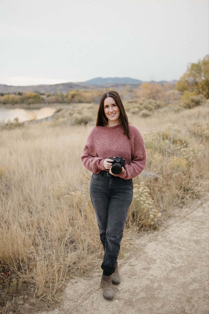 Holding Camera 桌布 壁紙 — 一位微笑的女子身穿粉紅色毛衣，沿著一條泥路穿過乾草，背景有山脈和湖泊。