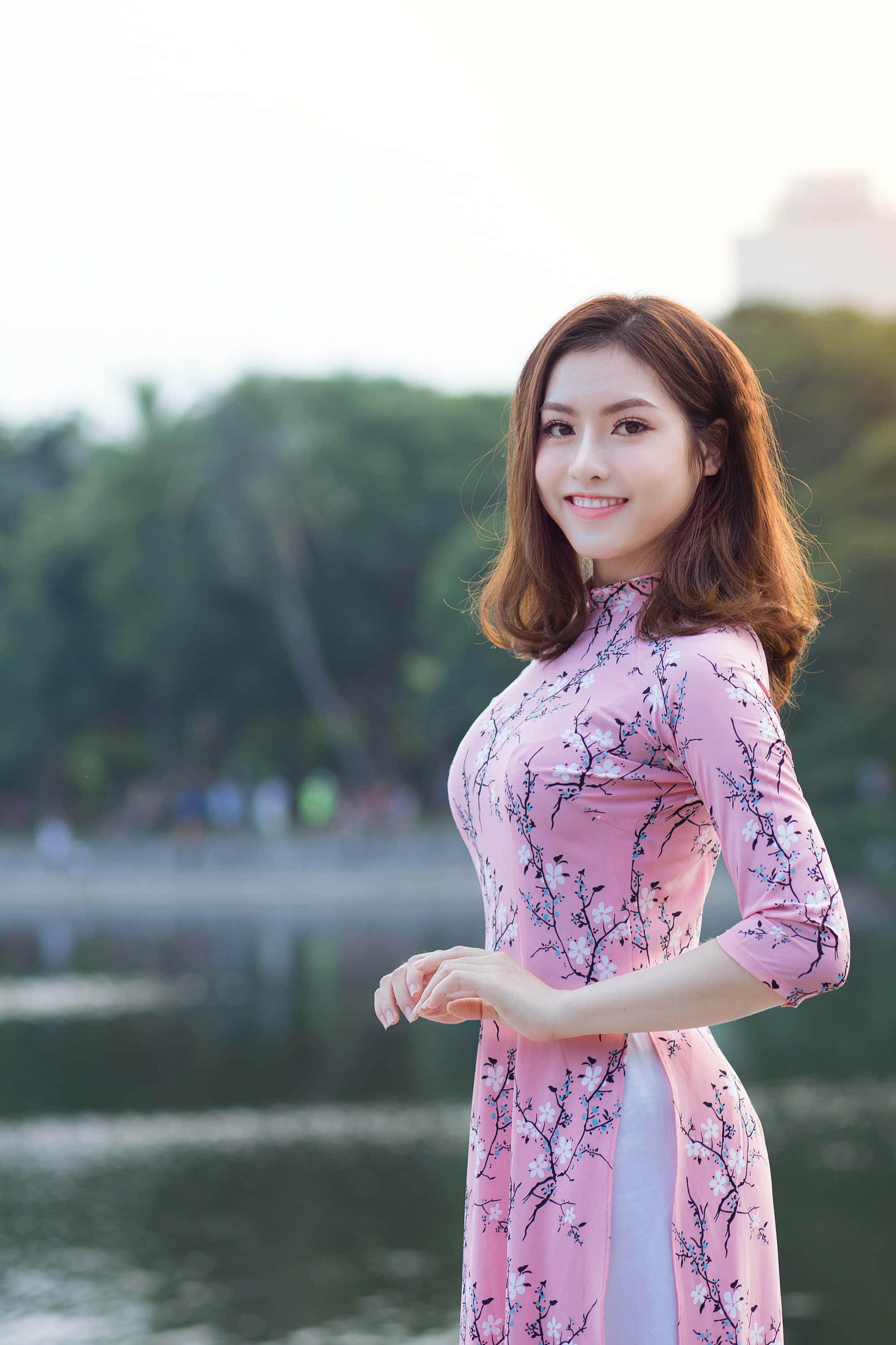 美女 桌布 壁紙 — 一位微笑的女子身穿粉紅花卉奧黛，優雅地站在寧靜的湖邊，身後樹木呈現柔焦效果。