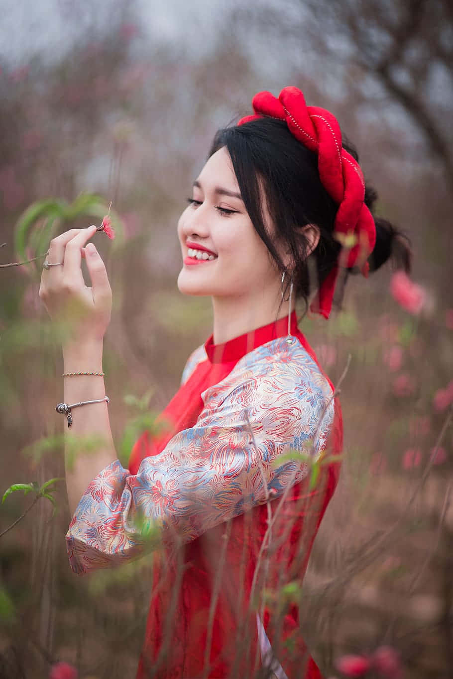 時尚 桌布 壁紙 — 一位微笑的女子身穿紅色圖案連衣裙和相配的頭飾，在柔和的春日花園中輕輕捧著一朵櫻花。