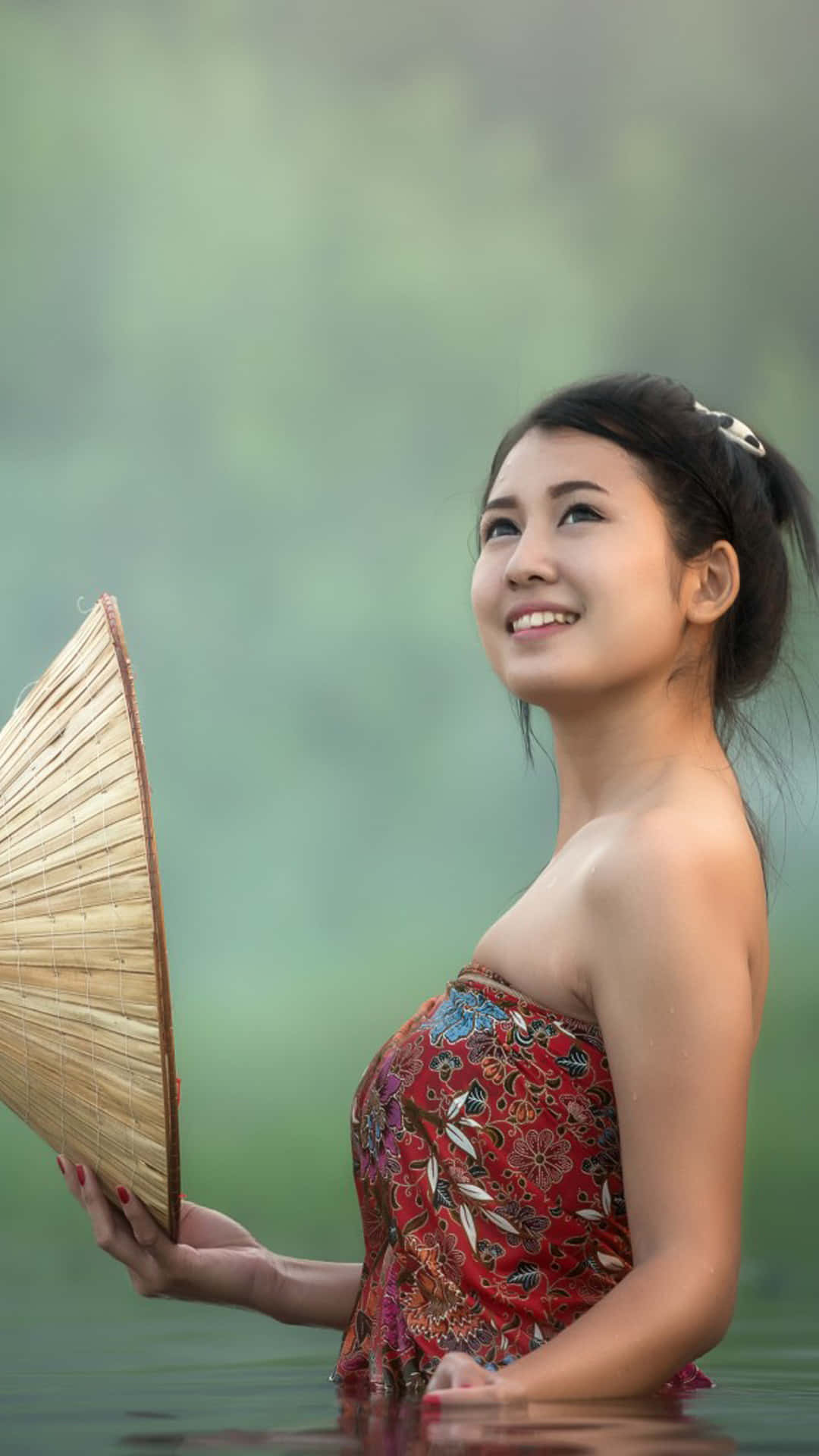 Asian Woman 桌布 壁紙 — 一位微笑的女子身穿紅色圖案連衣裙，手持竹扇，站在平靜的水中，背景是柔和的綠色迷霧。