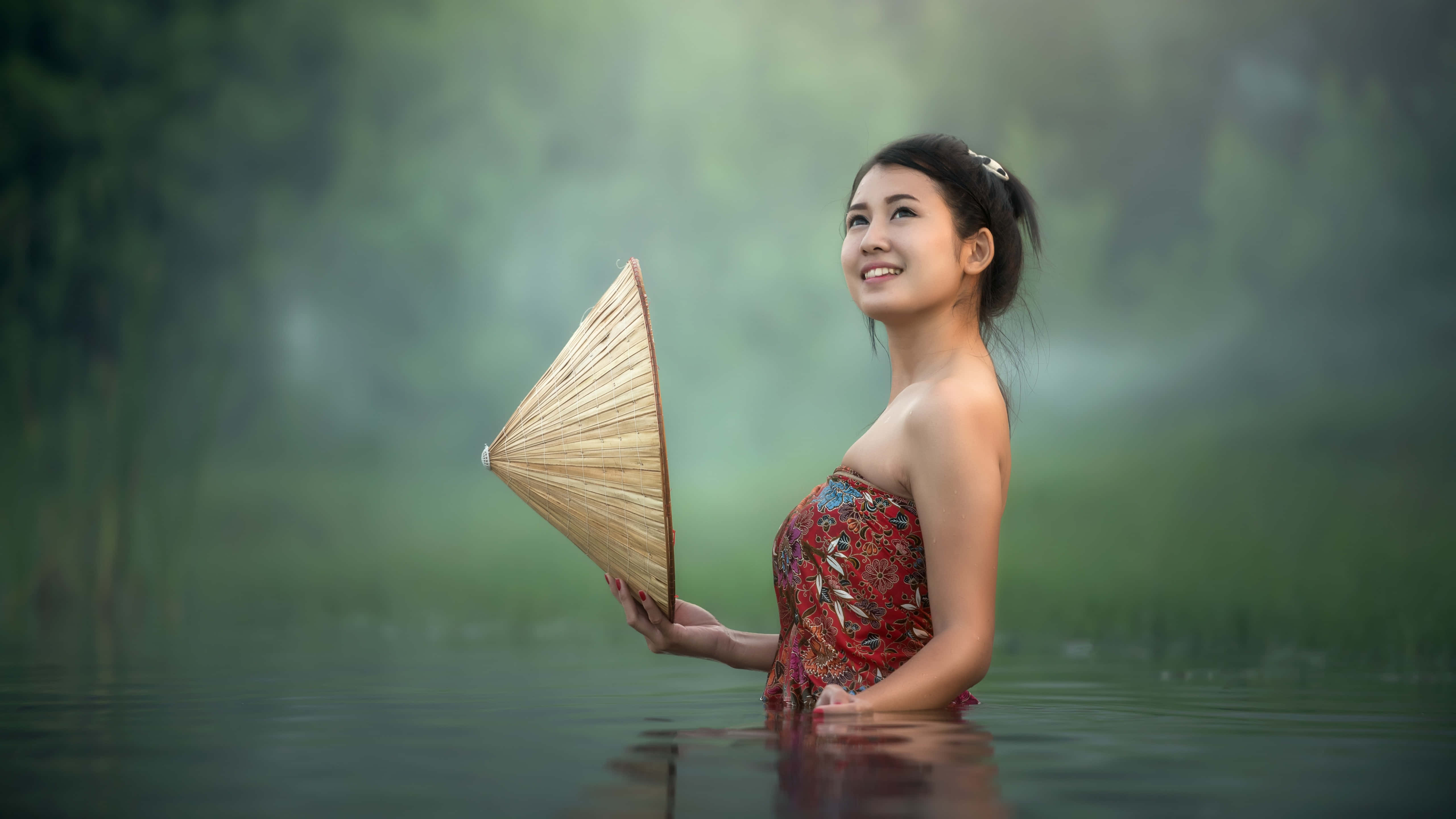 美女 桌布 壁紙 — 一位微笑的女子身穿紅色圖案連衣裙，手持草帽，站在霧氣瀰漫的綠色水中，周圍是模糊的樹葉。