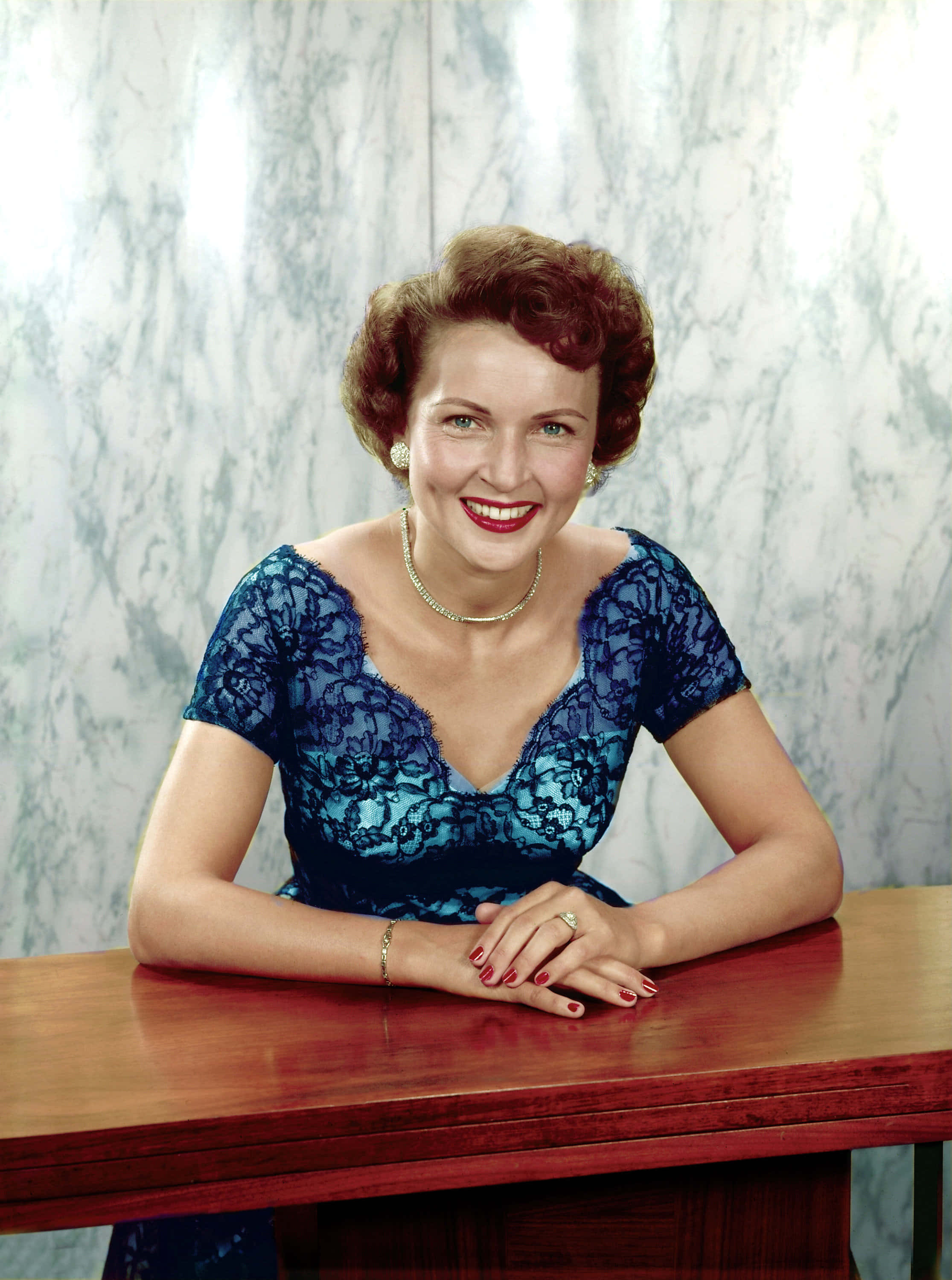 Betty White 桌布 壁紙 — 一位微笑的女子身穿藍色蕾絲連衣裙，在大理石牆前的木桌上優雅地擺姿勢。