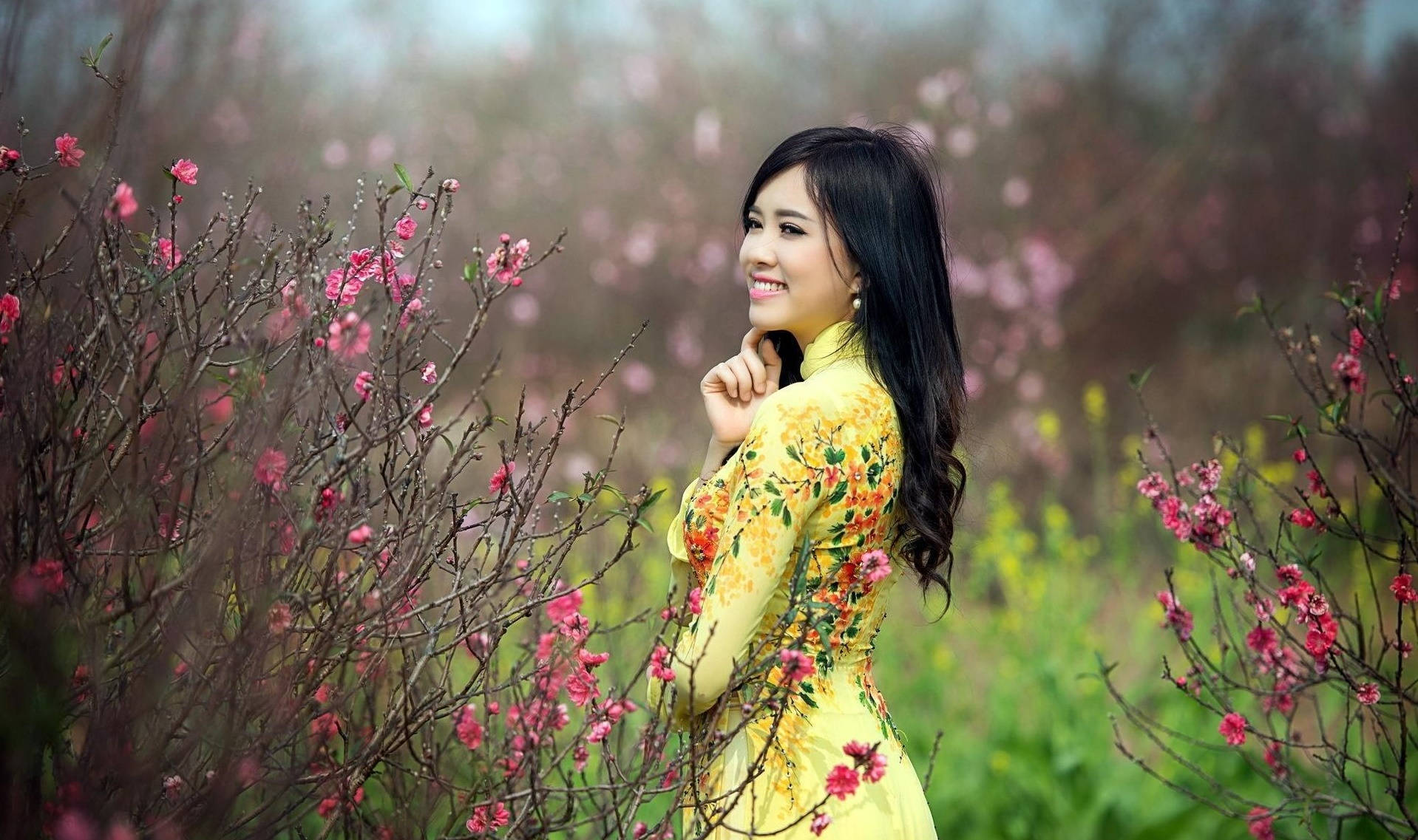 Woman 桌布 壁紙 — 一位微笑的女子身穿黃色花卉奧黛，站在柔焦花園中的粉紅花枝之間。