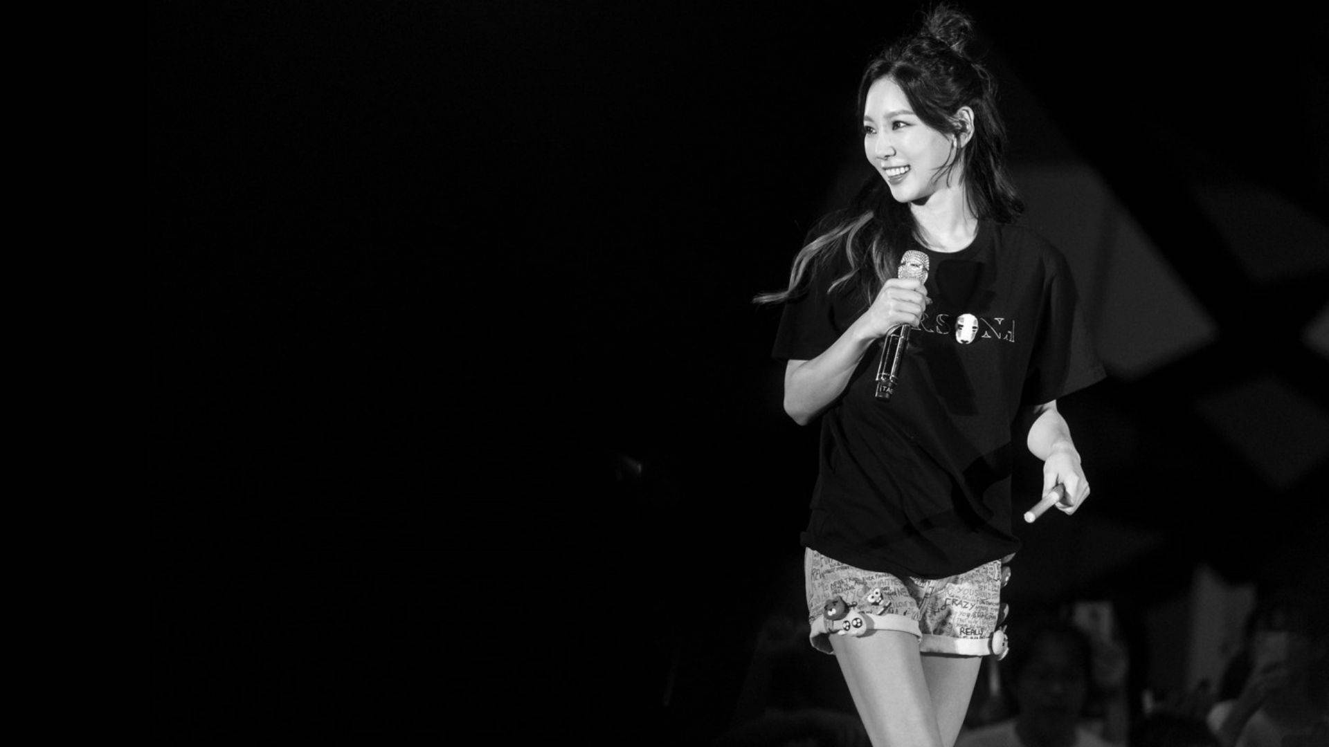 Taeyeon 桌布 壁紙 — 一位微笑的女子身穿黑色T恤和圖案短褲，在黑暗舞台的聚光燈下拿著麥克風。