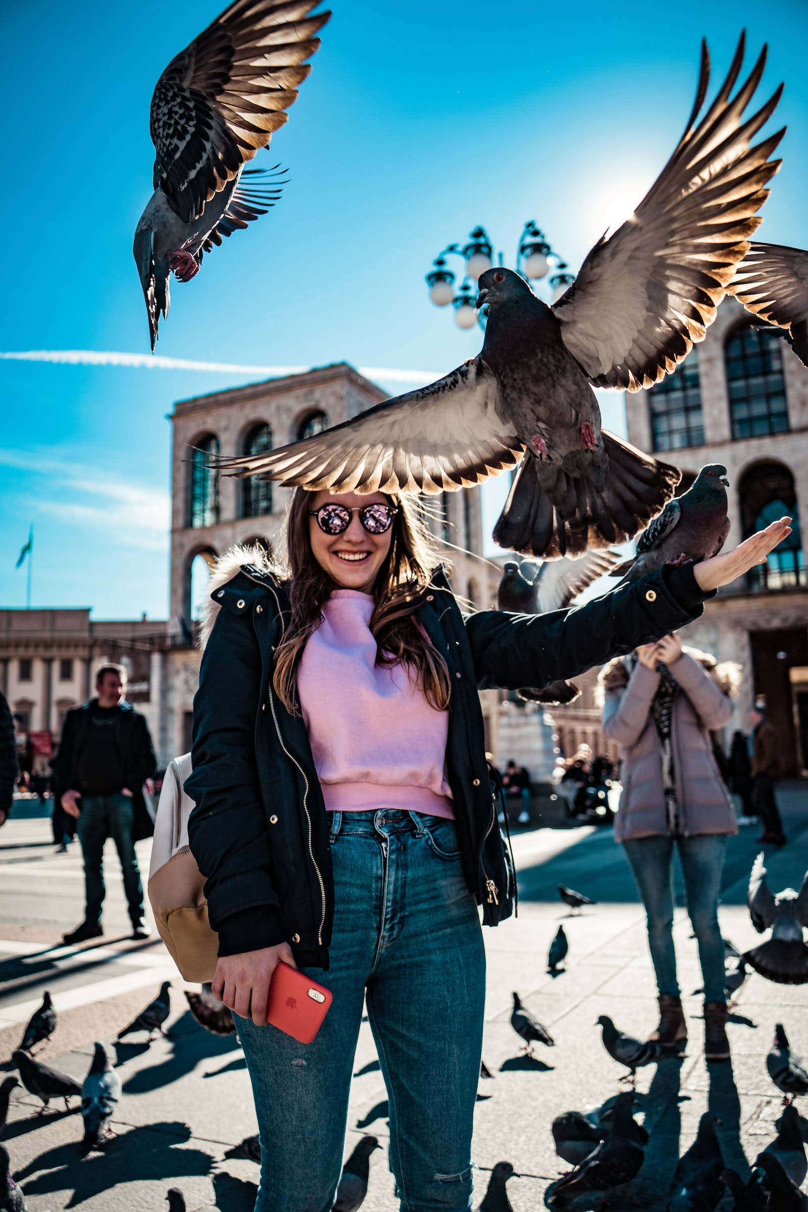 birds 桌布 壁紙 — 一位微笑的女子身穿黑色外套和粉紅色毛衣，在陽光明媚的城市廣場上，以明亮的藍天為背景，餵食展翅的鴿子。