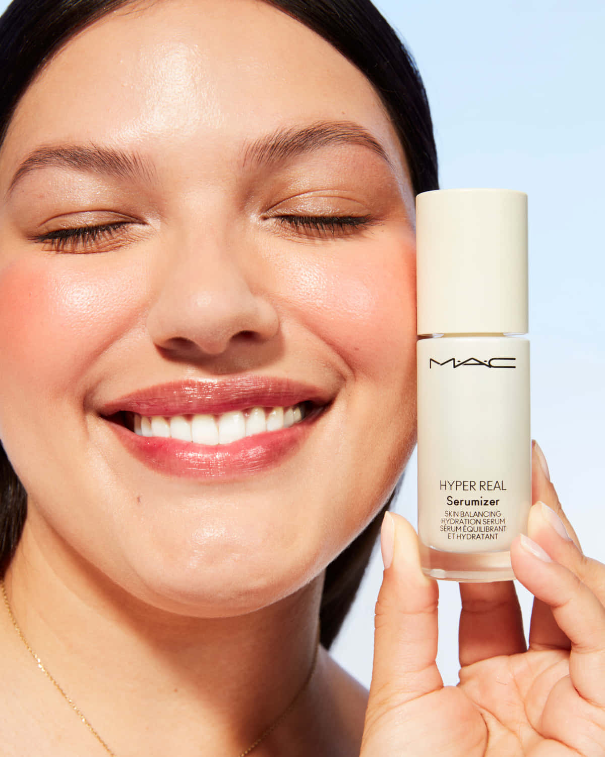 Mac Cosmetics 桌布 壁紙 — 一位微笑的女子閉著眼睛，手持MAC Hyper Real Serum瓶子，背景是柔和的藍色。