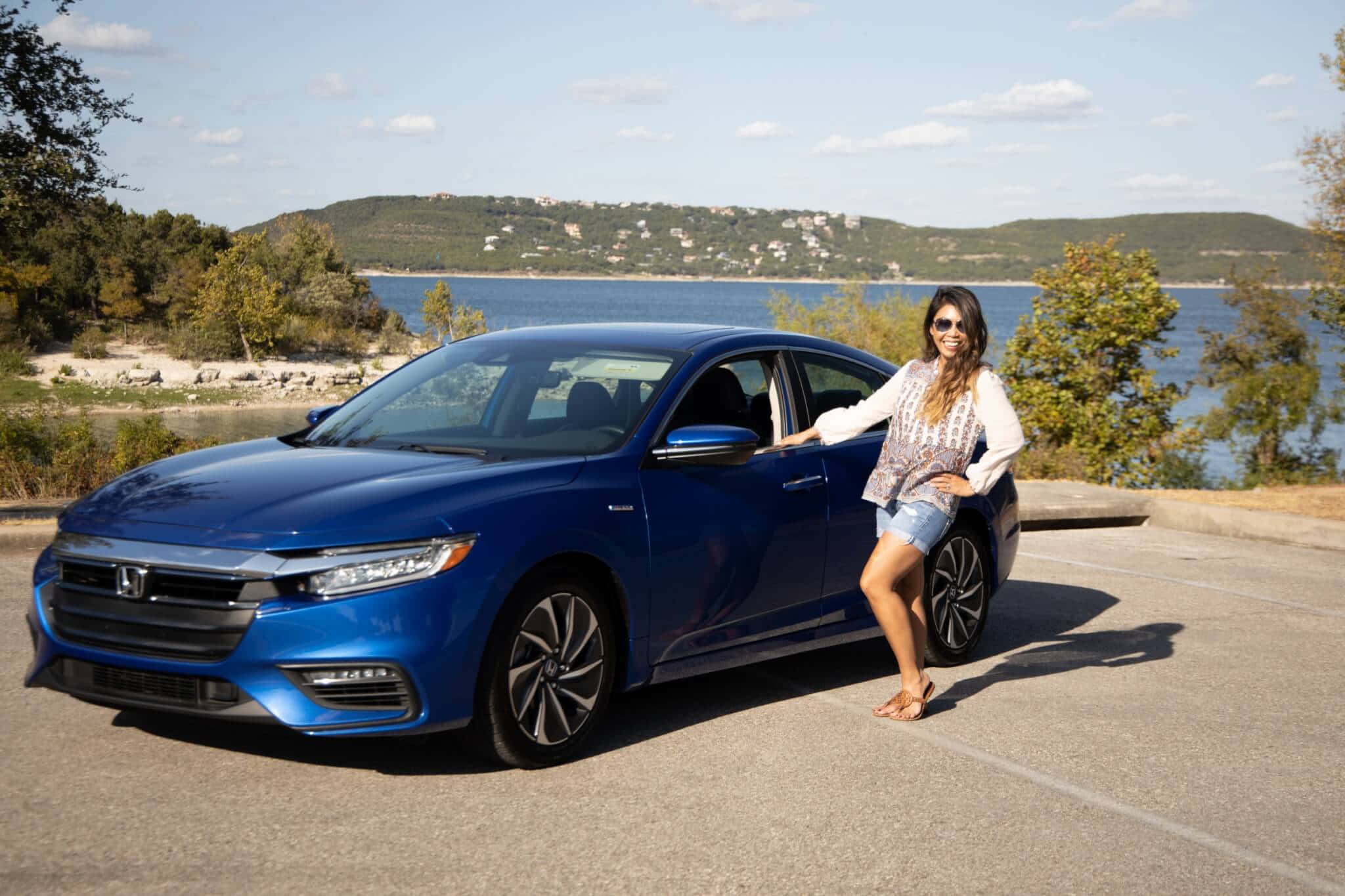 Honda Insight 桌布 壁紙 — 一位微笑的女子靠在一輛閃亮的藍色本田轎車旁，車停在風景如畫的湖邊，背景是起伏的丘陵。