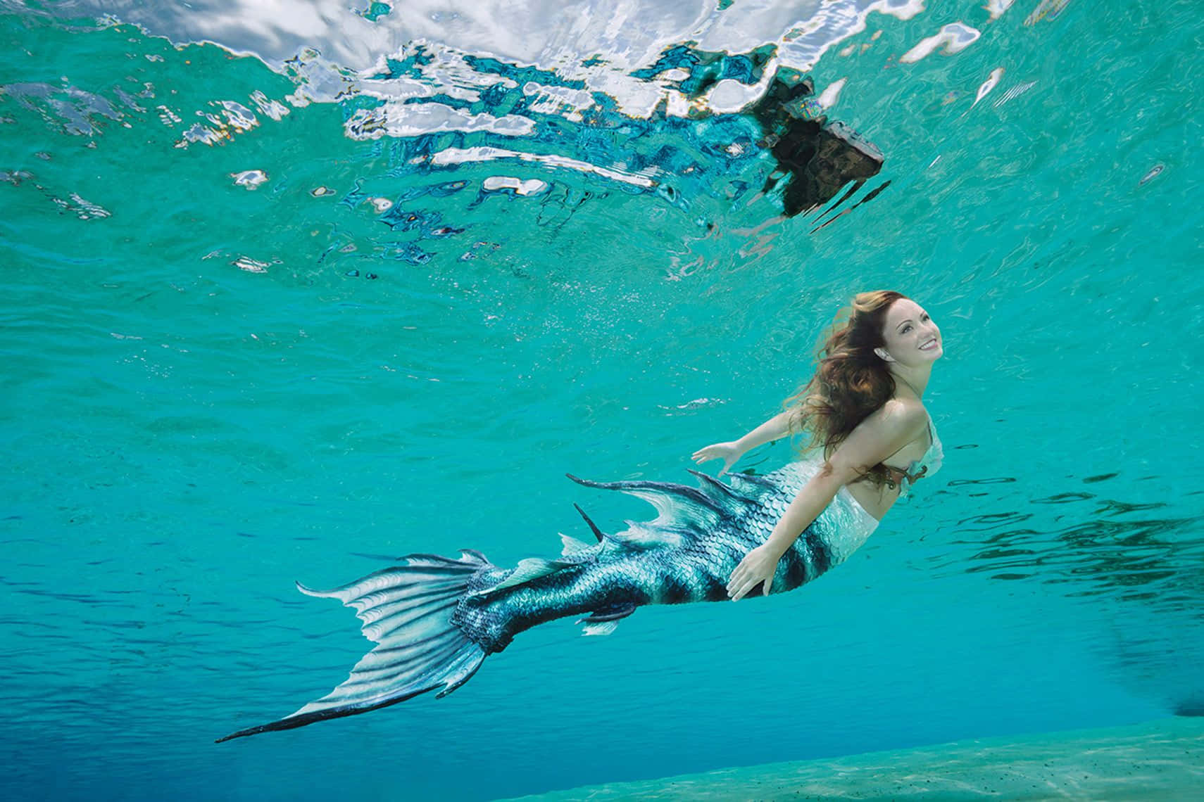 Mermaid Real Life 桌布 壁紙 — 一位微笑的女性，擁有藍色和銀色的人魚尾巴，在清澈的碧綠水中優雅地滑行。