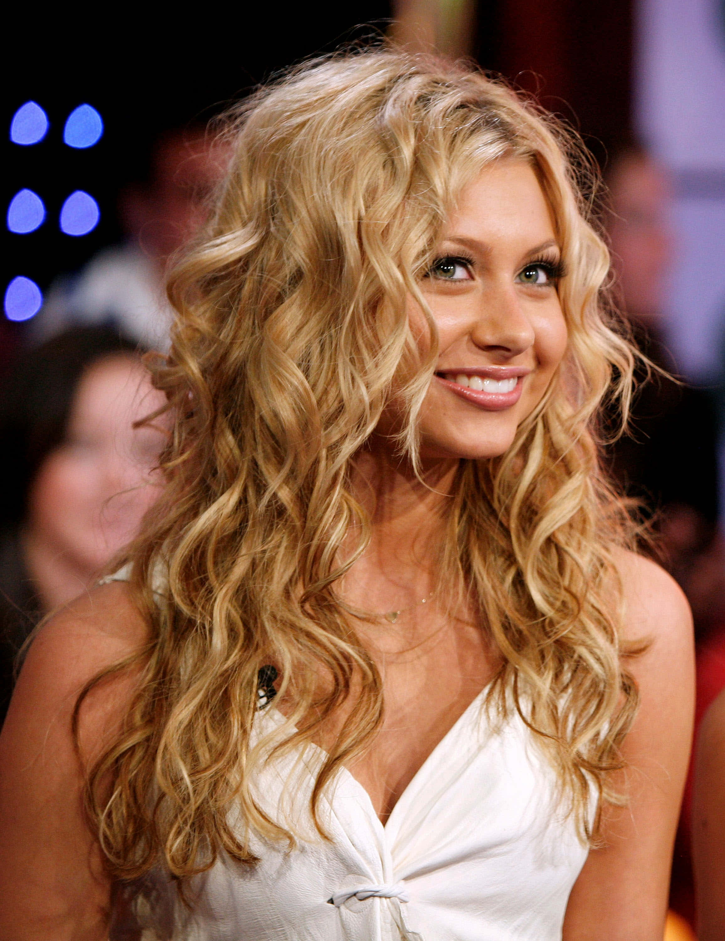 aly michalka 桌布 壁紙 — 一位微笑的女性，擁有豐盈的金色捲髮，穿著白色連衣裙，背景是模糊的活動場景，帶有藍色燈光。
