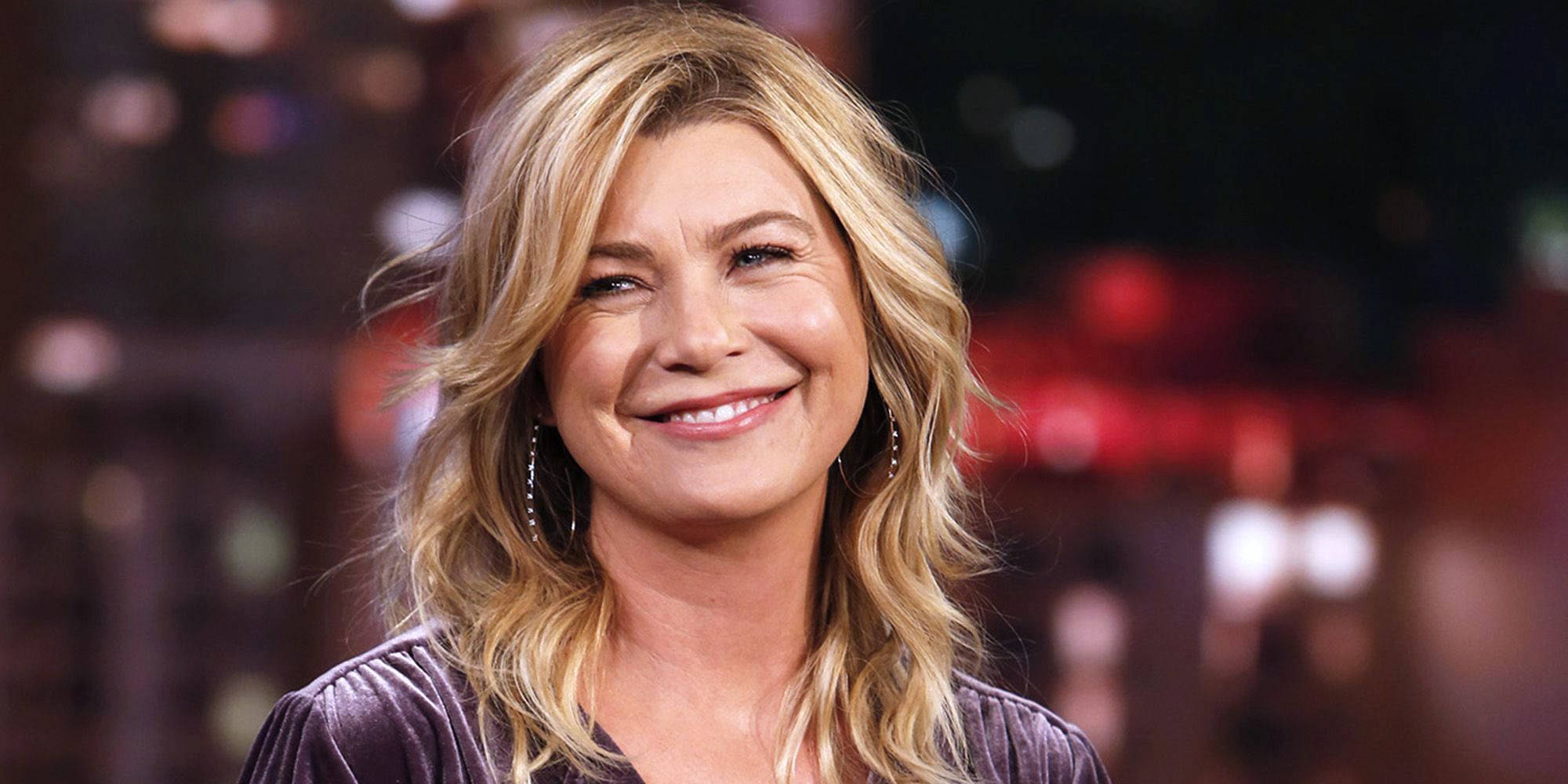 Ellen Pompeo 桌布 壁紙 — 一位微笑的女性，波浪金髮，戴著圓環耳環，穿著深色天鵝絨上衣，背景是模糊的城市夜景。