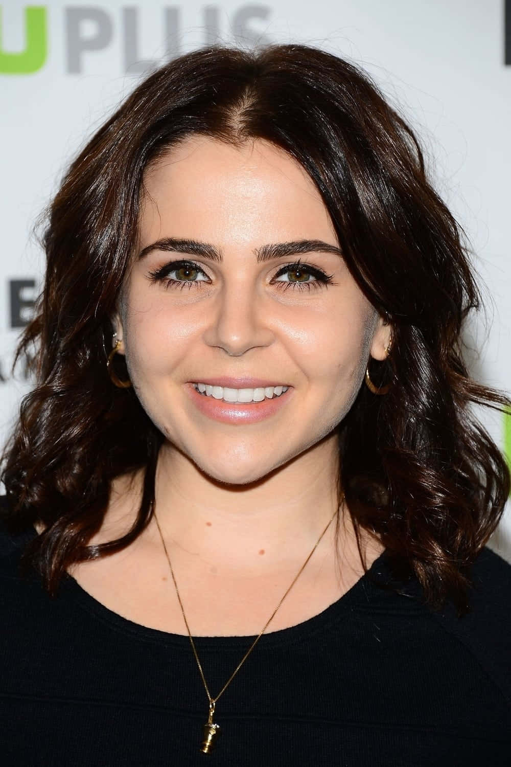 Mae Whitman 桌布 壁紙 — 一位微笑的女性，深色波浪長髮，戴著金色圓環耳環，在活動中穿著黑色上衣和項鍊。