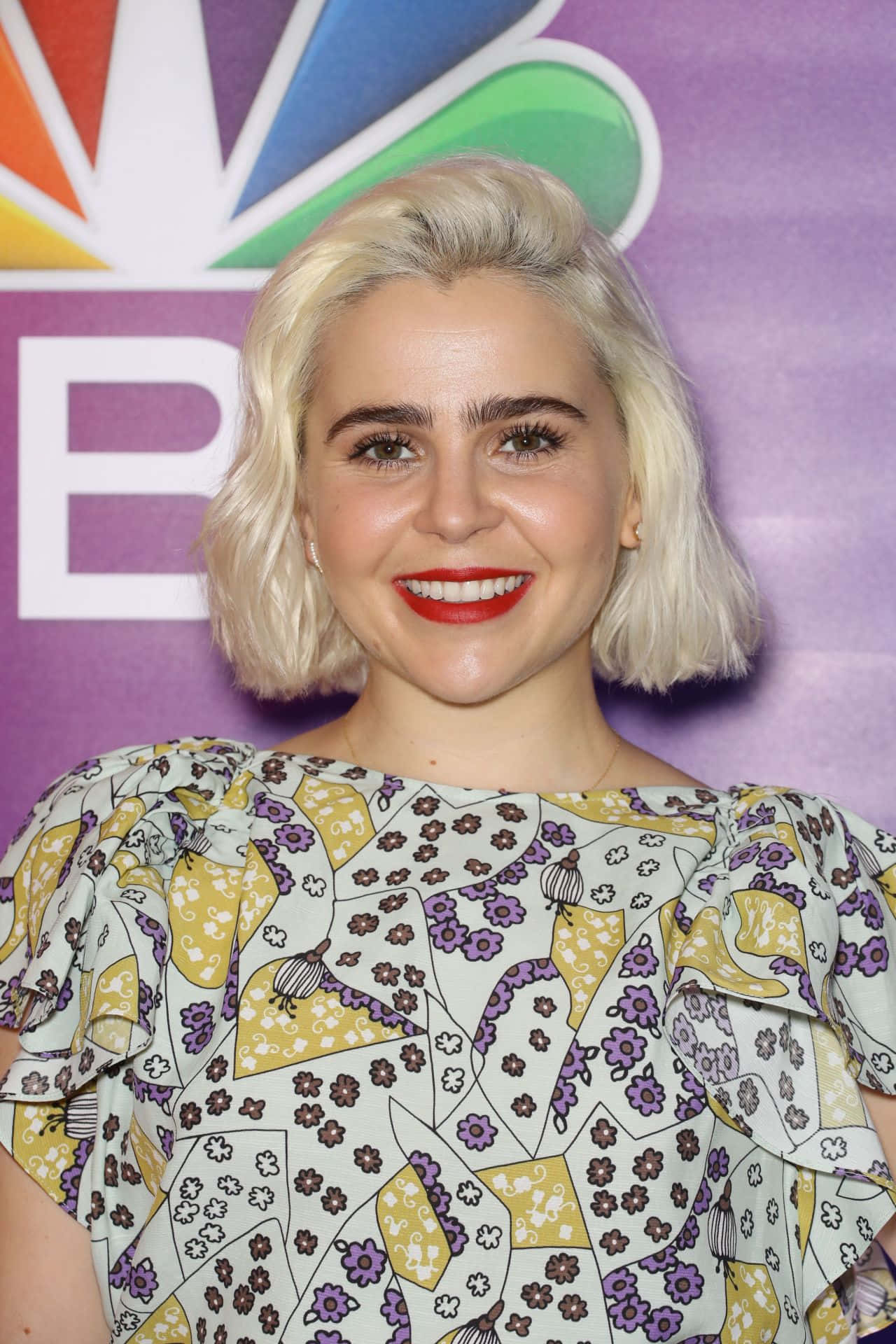 Mae Whitman 桌布 壁紙 — 一位微笑的女性，留著鉑金色鮑伯頭，塗著大膽的紅唇，身穿花卉圖案襯衫，背景是紫色的NBC。
