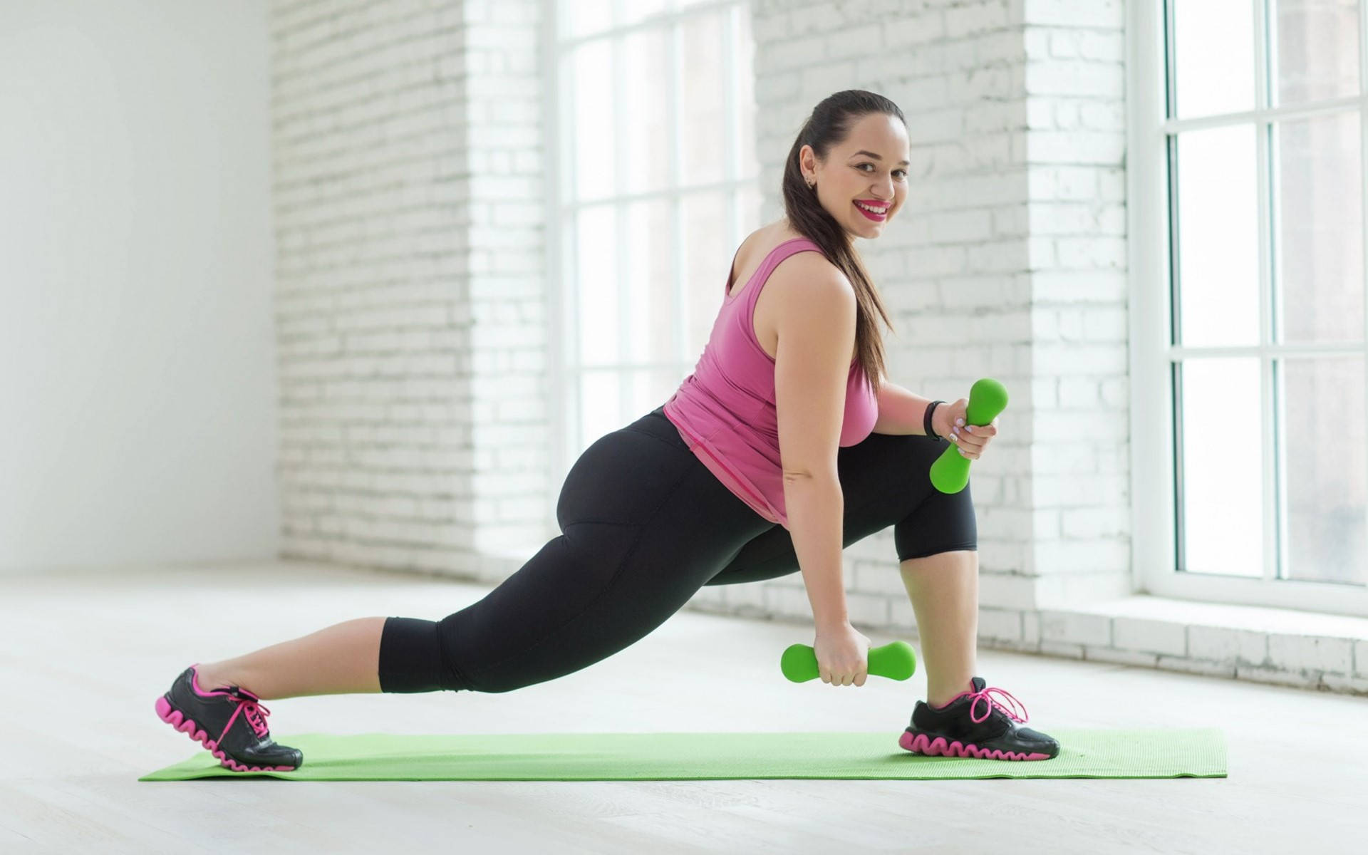 Exercise 桌布 壁紙 — 一位微笑的女性，身穿粉紅色上衣和黑色緊身褲，在明亮的白色磚房中的綠色墊子上用綠色啞鈴做弓步。