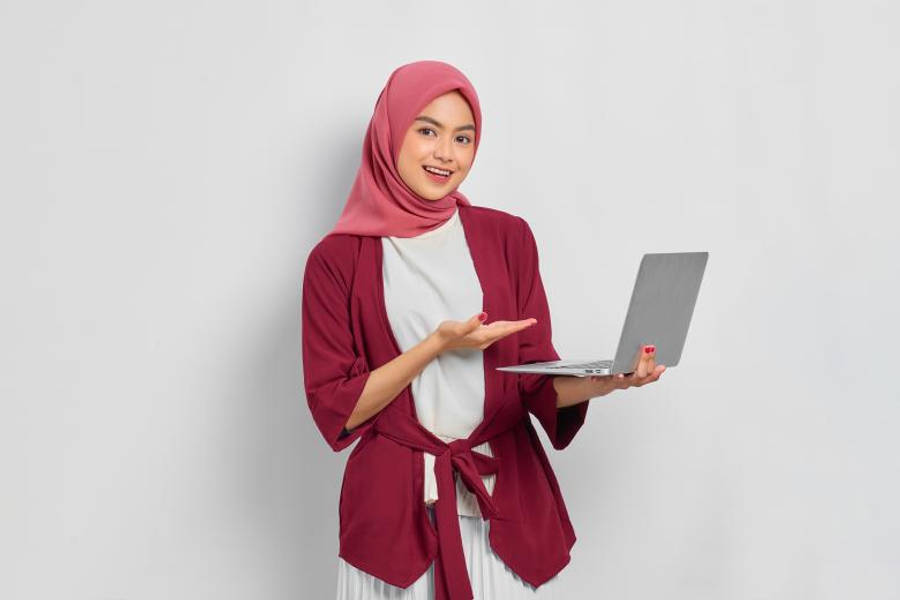 HD Islamic 桌布 壁紙 — 一位微笑的女性，身穿粉紅色頭巾和栗色長袍，在純白背景前拿著筆記型電腦，並熱情地做手勢。