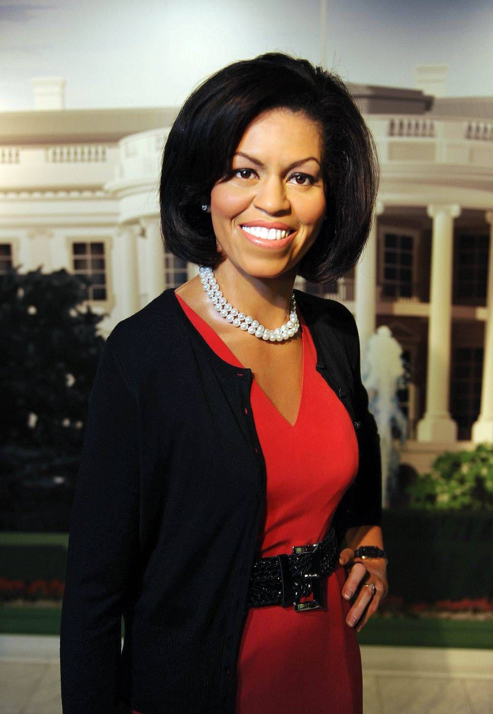 Michelle 桌布 壁紙 — 一位微笑的女性，身穿紅色連衣裙和黑色開襟衫，戴著珍珠項鍊，站在白宮背景前。