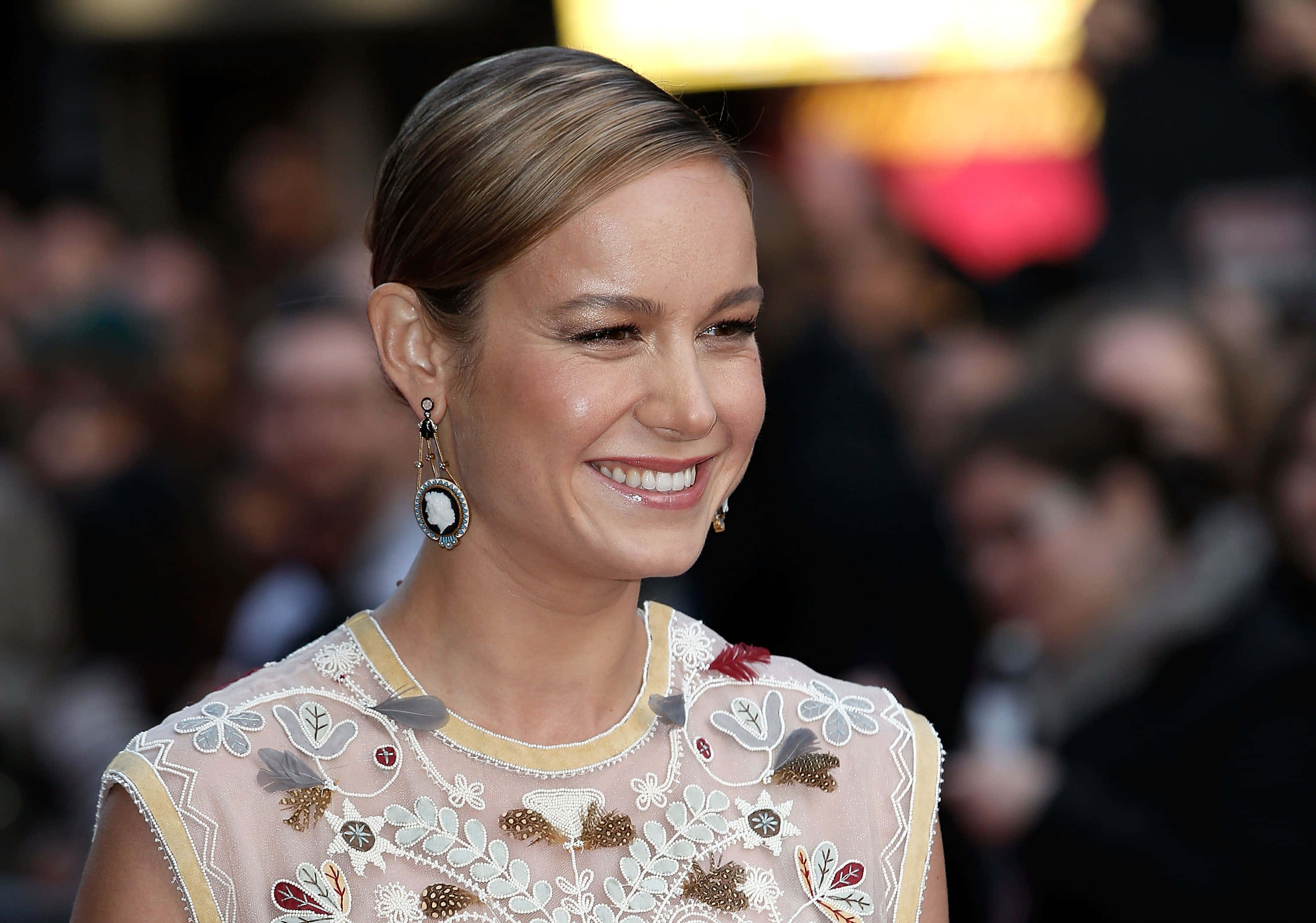 brie larson 桌布 壁紙 — 一位微笑的女性，身穿華麗刺繡連衣裙，佩戴浮雕耳環，身處擁擠的活動中。