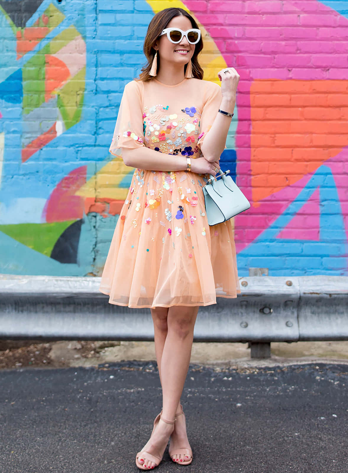 Spring Fashion 桌布 壁紙 — 一位微笑的女性，身穿蜜桃色花卉洋裝和白色太陽眼鏡，自信地站在充滿活力的塗鴉磚牆前。