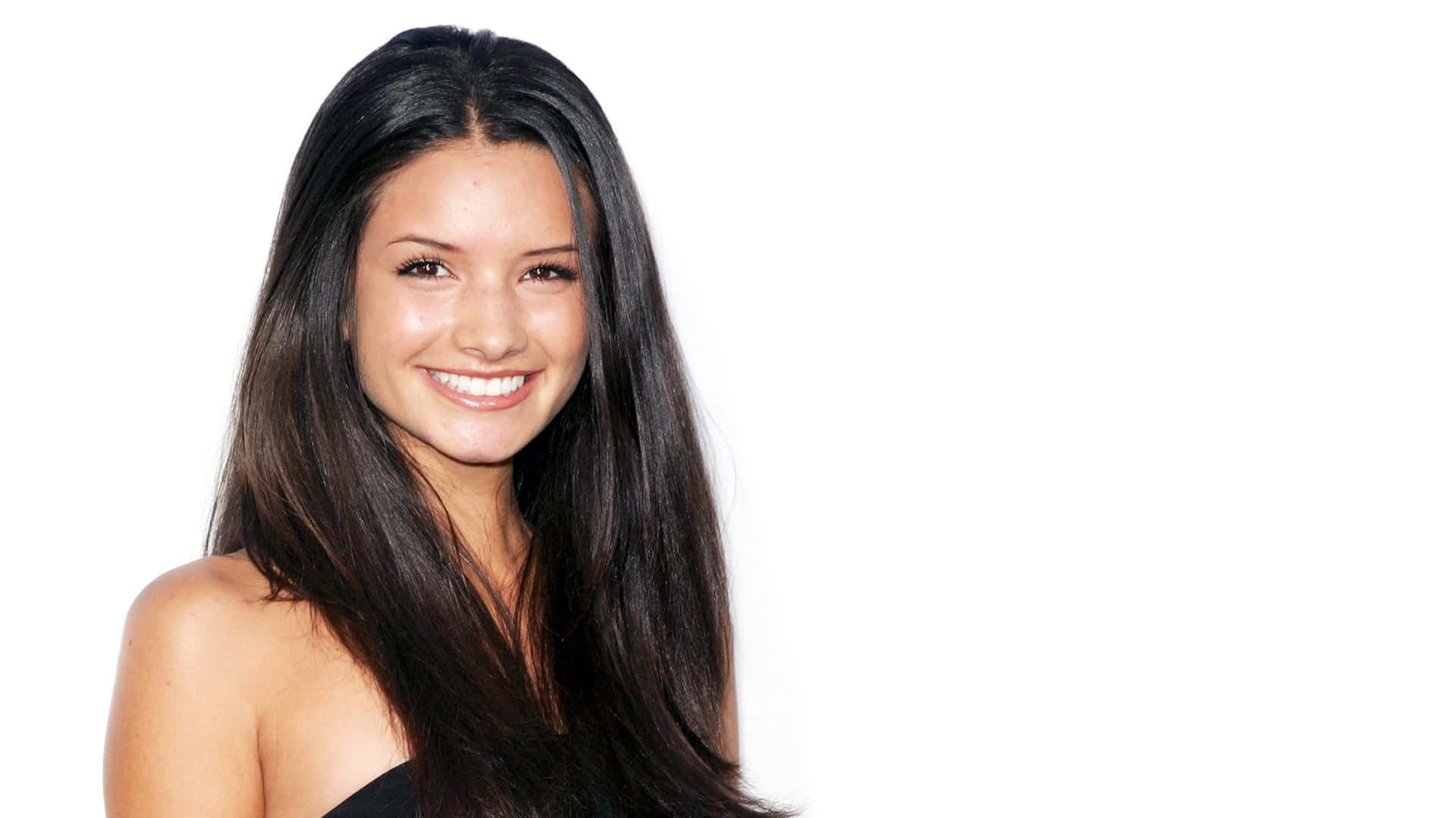 Alice Greczyn 桌布 壁紙 — 一位微笑的女性，長長的黑髮，穿著黑色上衣，背景是純白色。
