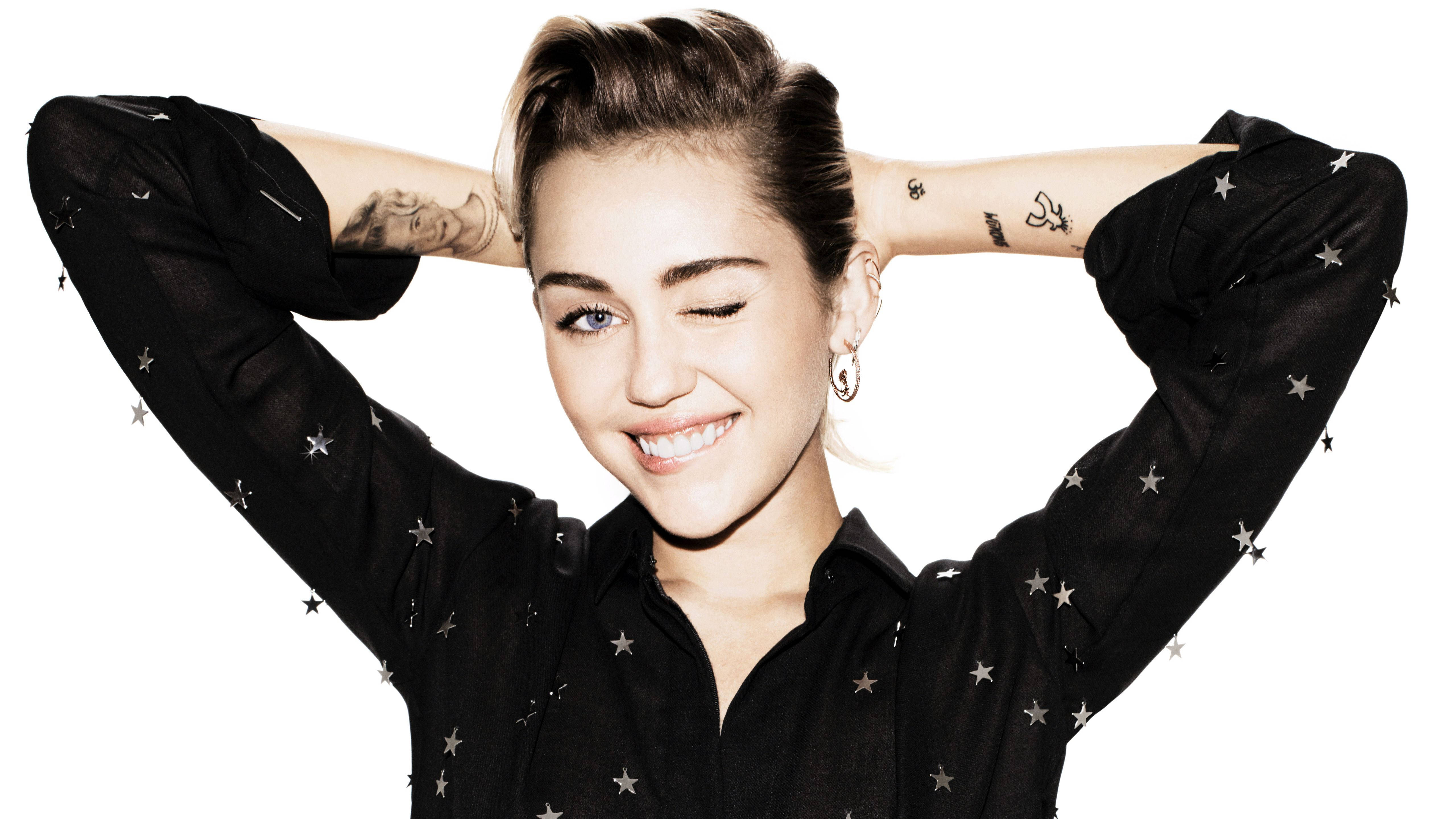 miley 桌布 壁紙 — 一位微笑的女性，閉上一隻眼，雙手舉過頭頂，身穿黑色星星圖案襯衫，背景為白色。