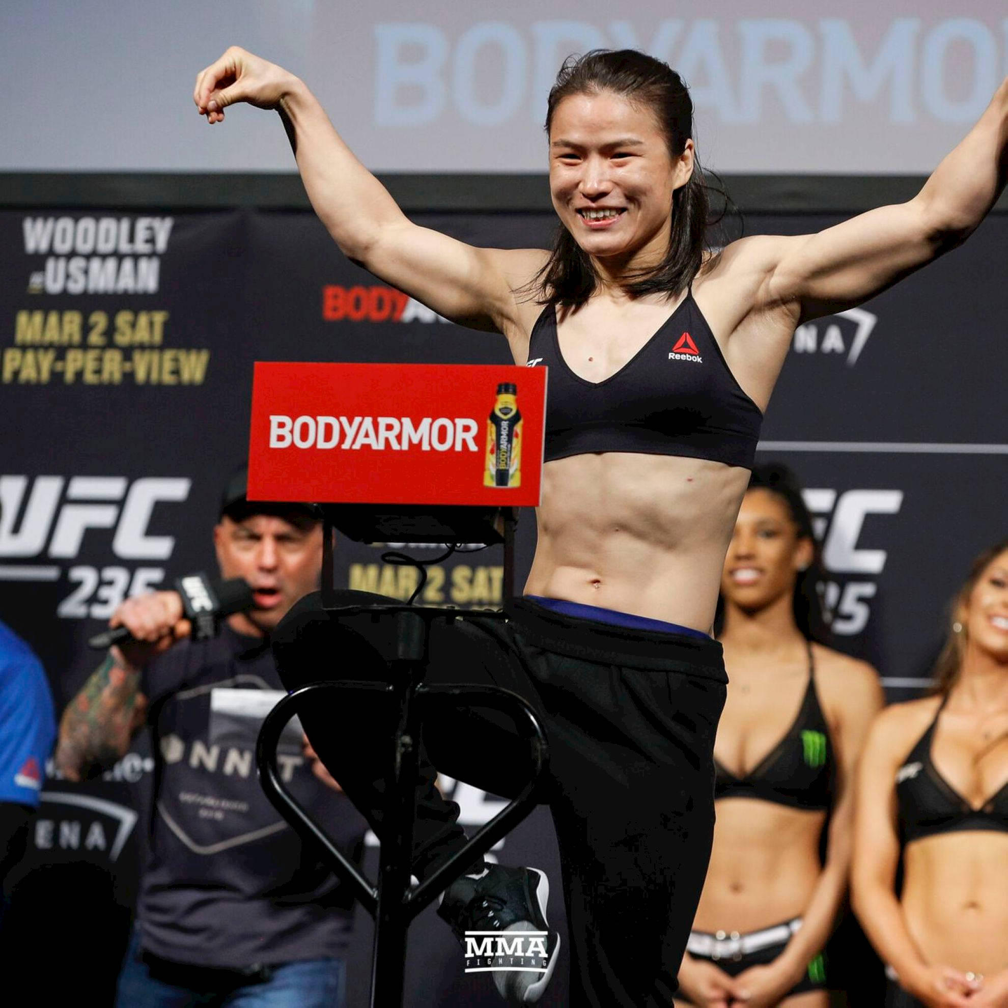 Athletic 桌布 壁紙 — 一位微笑的女性MMA拳手在UFC過磅活動中，在磅秤上展示肌肉，舞台燈光璀璨。
