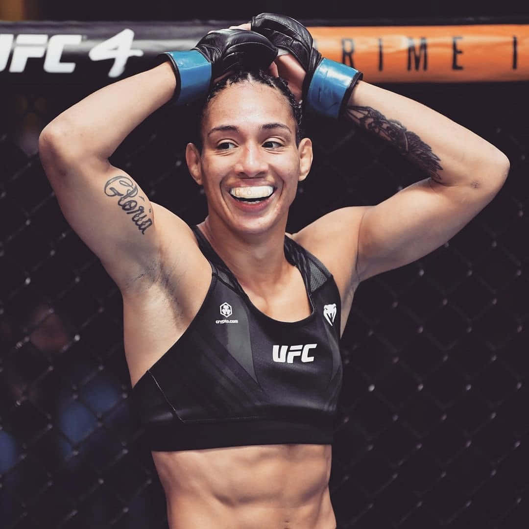 UFC 桌布 壁紙 — 一位微笑的女性MMA拳手身穿黑色UFC運動內衣，在八角籠內慶祝，手臂有刺青，戴著藍色手套。