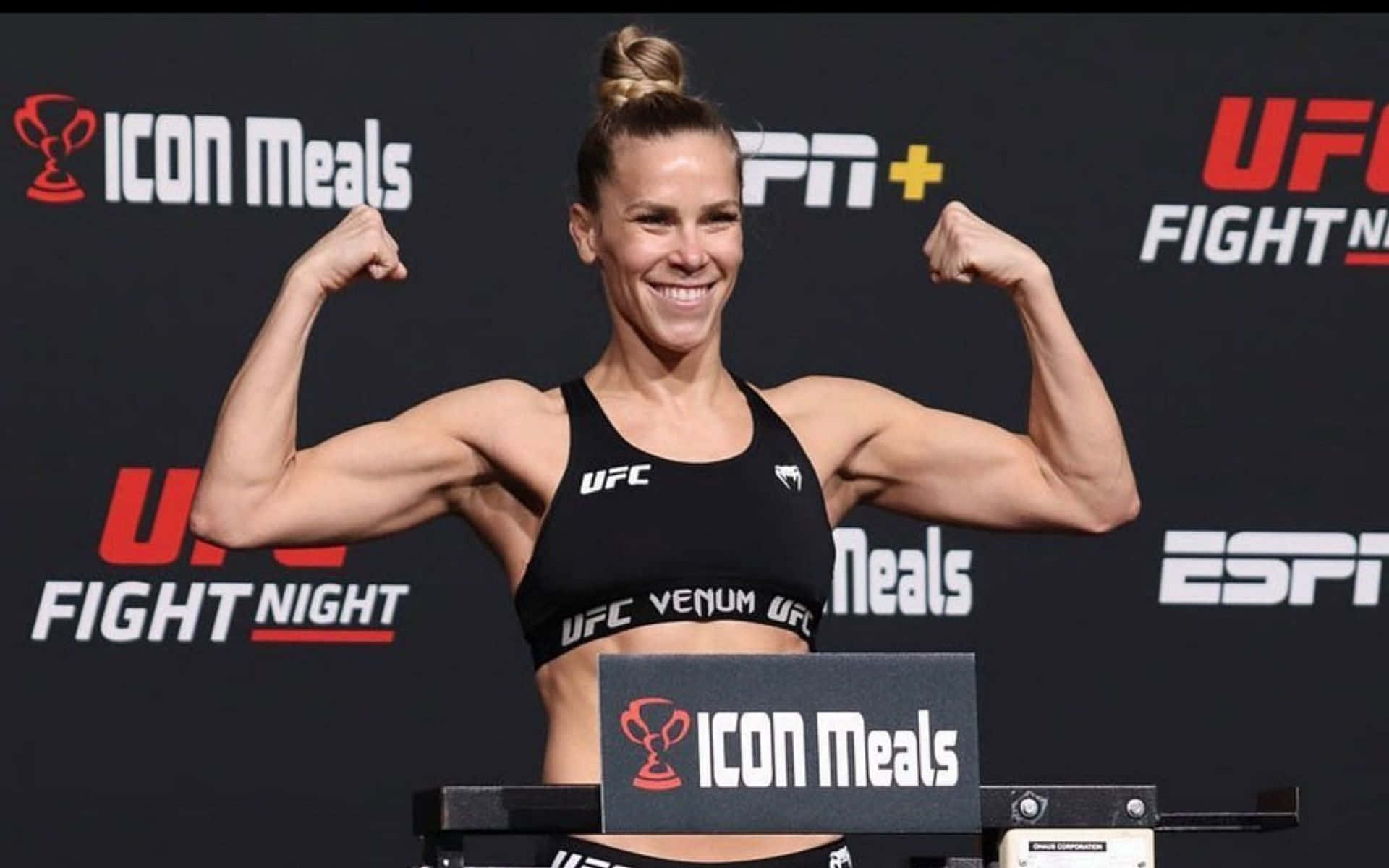 桌布 壁紙 — 一位微笑的女性UFC拳手在有品牌背景的稱重活動中彎曲她肌肉發達的手臂。