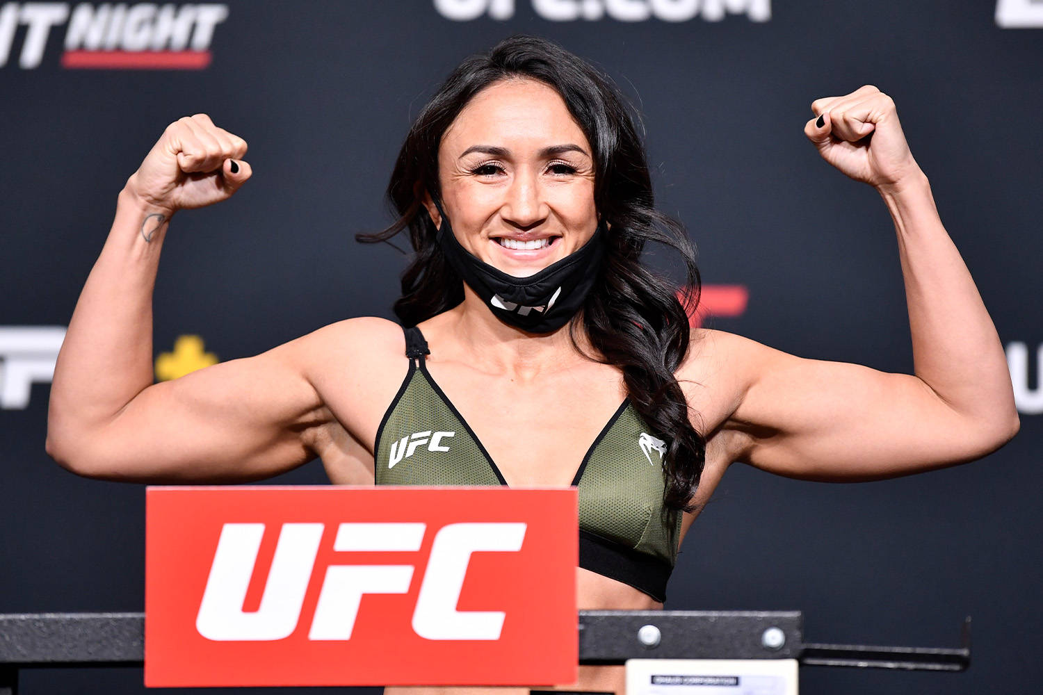 MMA 桌布 壁紙 — 一位微笑的女性UFC拳手在稱重活動中展示肌肉，穿著綠色運動胸罩和黑色口罩。