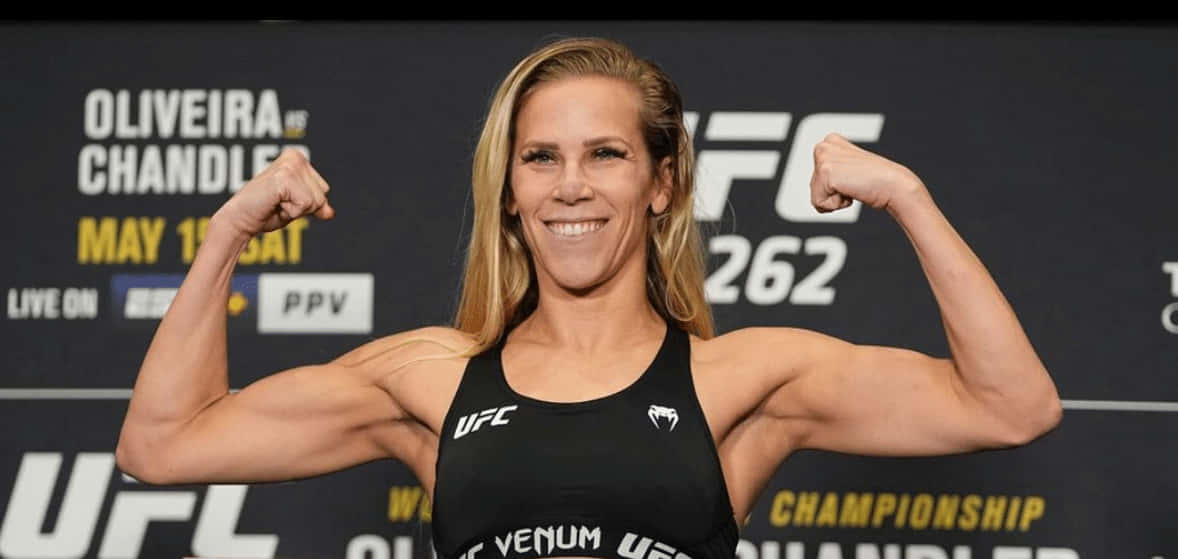 桌布 壁紙 — 一位微笑的女性UFC拳手在稱重活動中彎曲她肌肉發達的手臂，身後有冠軍品牌標誌。