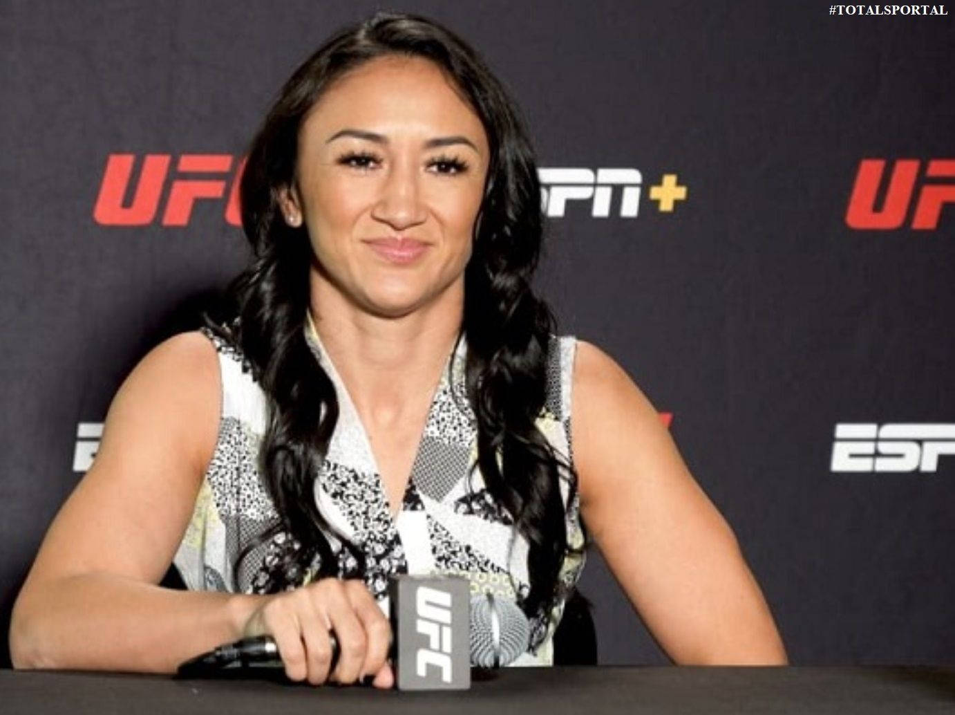 MMA 桌布 壁紙 — 一位微笑的女性UFC拳手坐在記者會桌前，手持帶有UFC品牌標誌的麥克風。