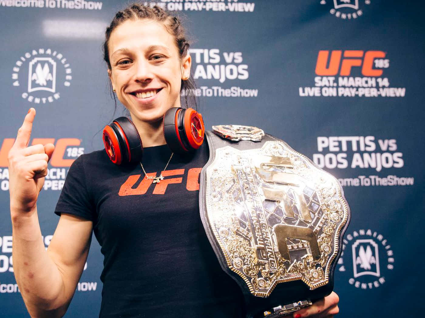 Champion 桌布 壁紙 — 一位微笑的女性UFC拳手自豪地舉起冠軍腰帶，戴著紅色耳機，同時向上指。