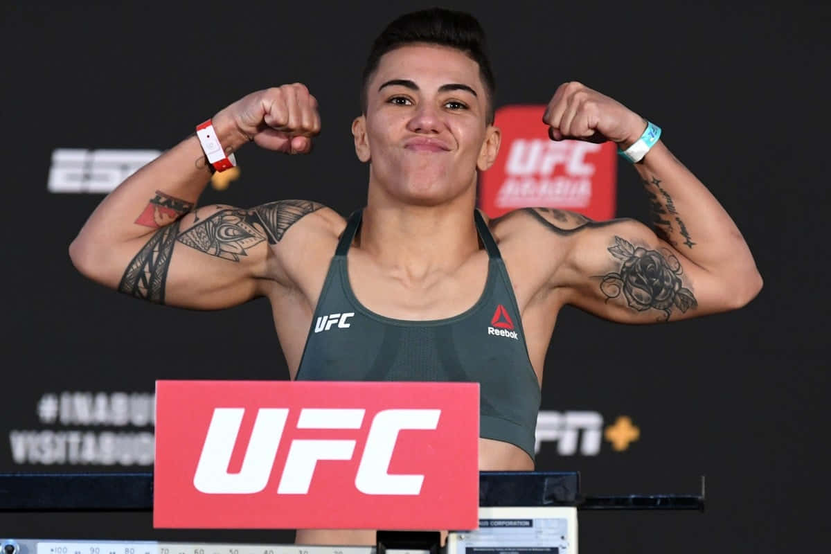 MMA 桌布 壁紙 — 一位微笑的女性UFC拳擊手在稱重活動中彎曲她帶有刺青的手臂，身後有品牌標誌。