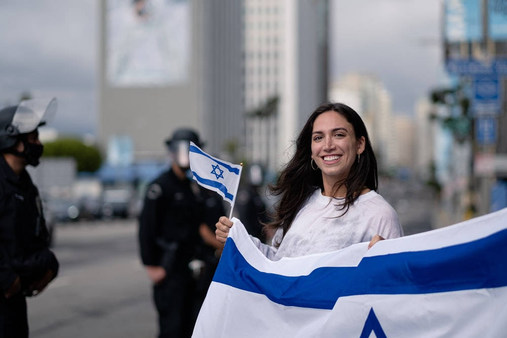 Israel Flag 桌布 壁紙 — 一位微笑的女性在城市的街道上自豪地舉著一面大型以色列國旗，背景中有警察站立。