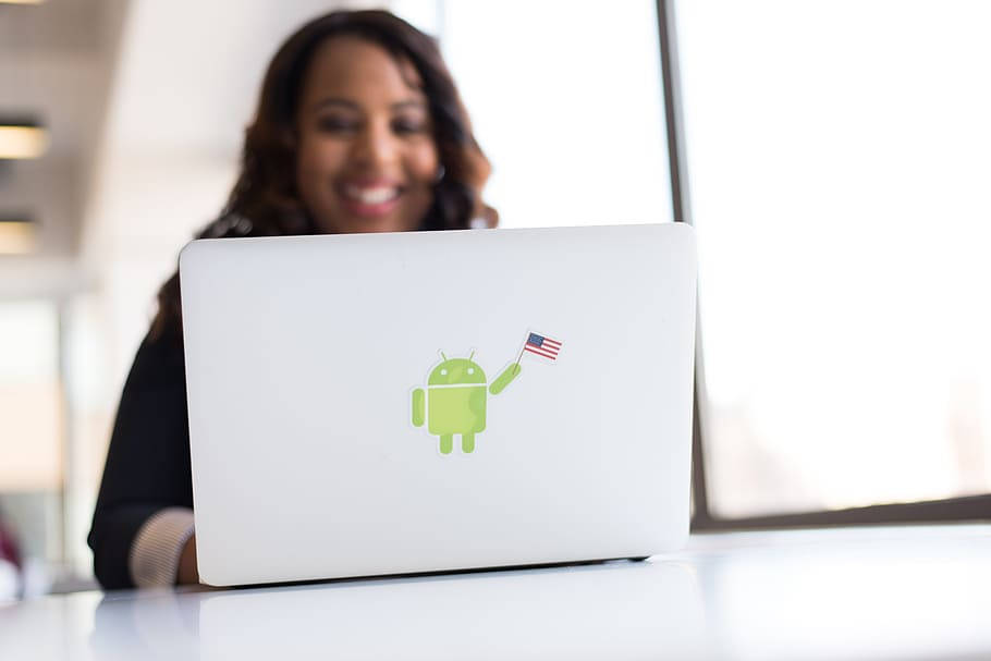 Android Developer 桌布 壁紙 — 一位微笑的女性坐在白色筆記型電腦後面，綠色Android吉祥物手持美國國旗貼紙。