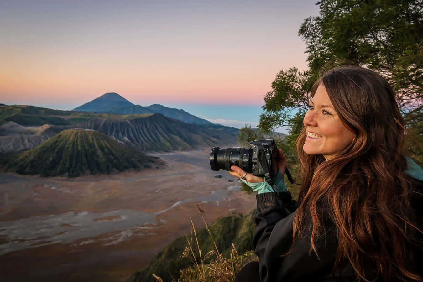 印尼 桌布 壁紙 — 一位微笑的女性手持相機，捕捉火山地貌上層巒疊嶂的壯麗日出。