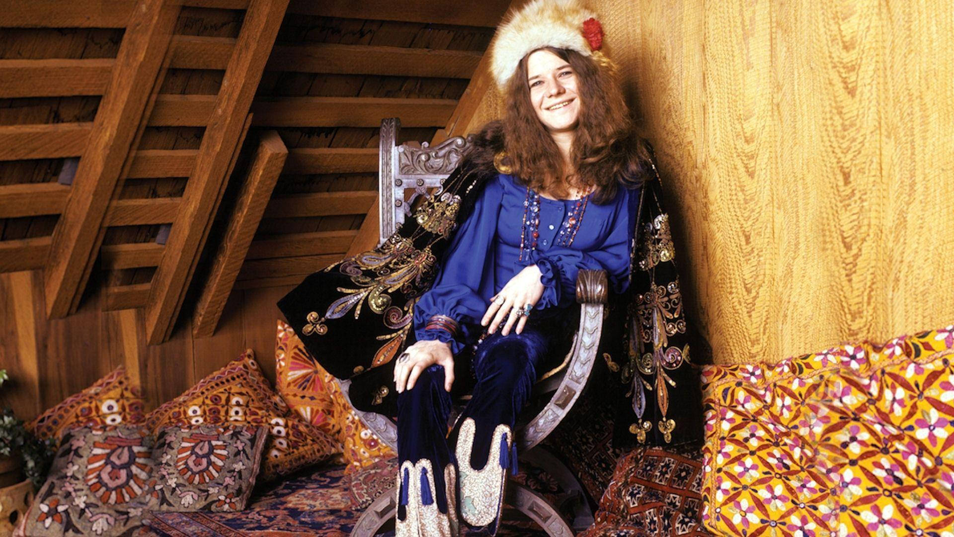 Entertainment 桌布 壁紙 — 一位微笑的女性穿著1970年代波希米亞風格服裝，優雅地坐在裝飾椅上，周圍是木製閣樓房間裡的圖案靠墊。