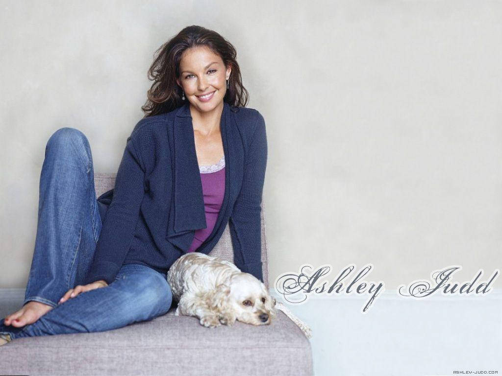 Ashley Judd 桌布 壁紙 — 一位微笑的女性穿著休閒服裝，放鬆地坐在沙發上，一隻毛茸茸的小狗依偎在她身旁休息。