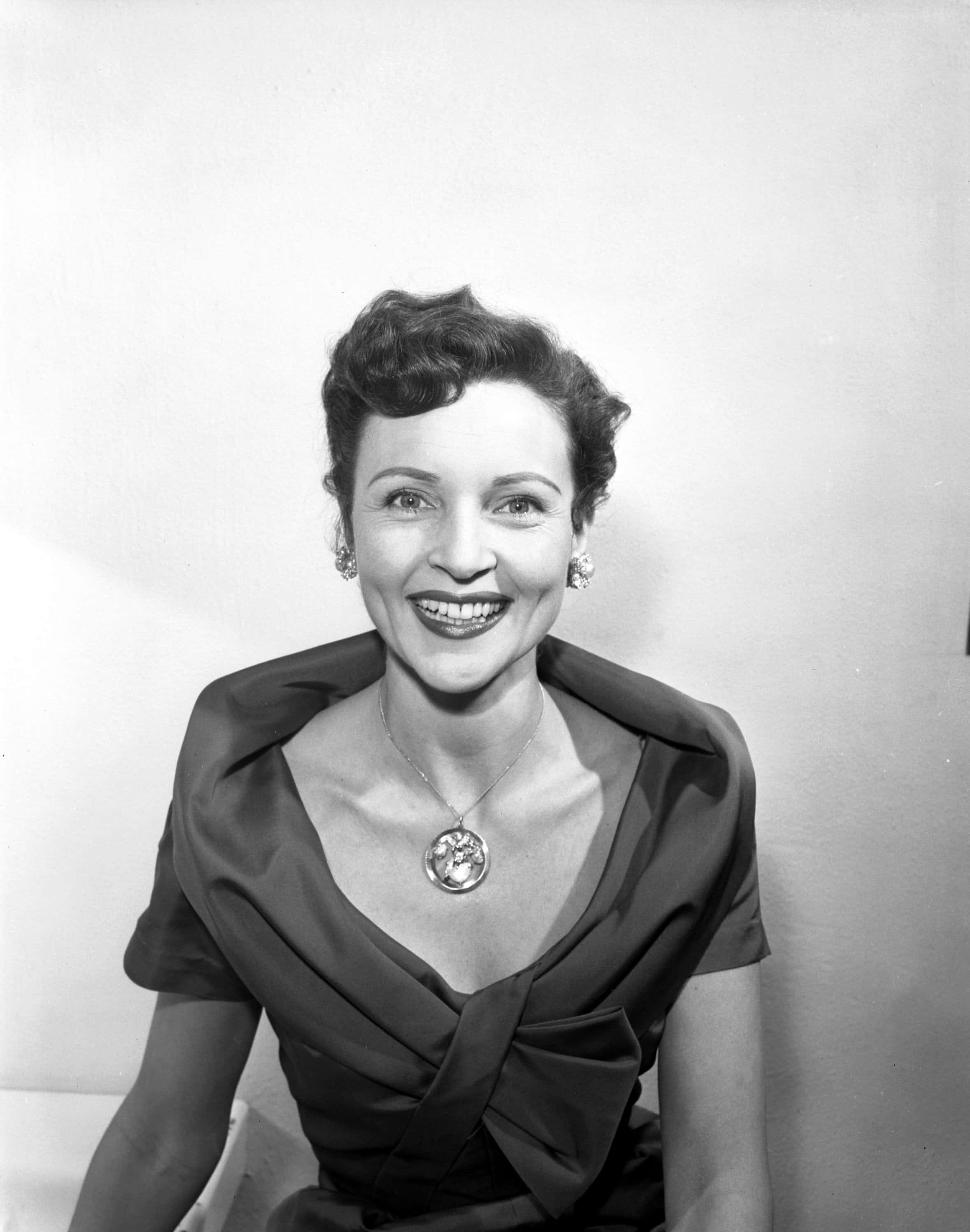 Betty White 桌布 壁紙 — 一位微笑的女性穿著時尚的1940年代連衣裙，戴著顯眼的項鍊和耳環，在純色背景前擺姿勢。