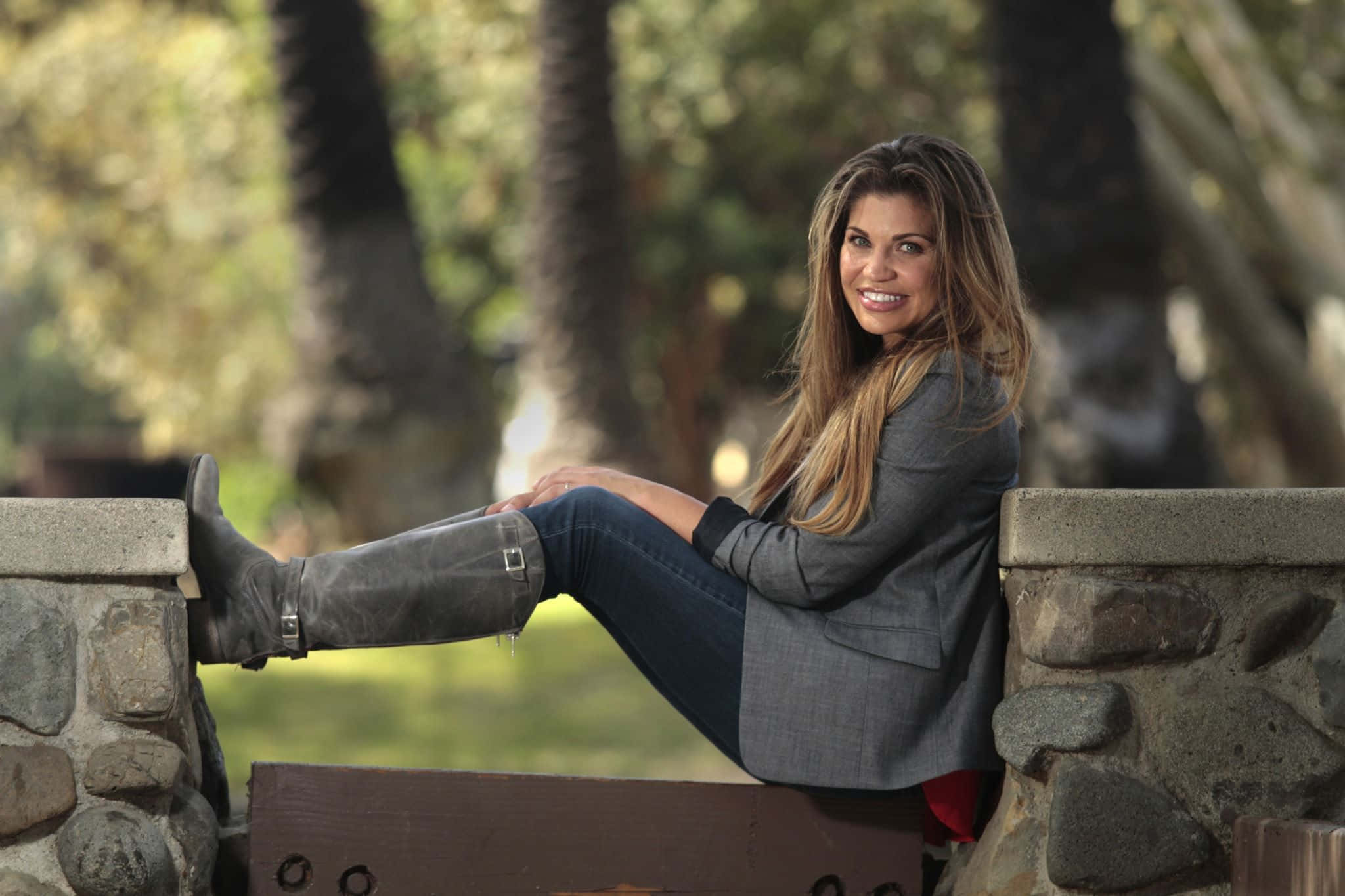 Danielle Fishel 桌布 壁紙 — 一位微笑的女性穿著灰色西裝外套和靴子，隨意地靠在一道石牆上，背後的棕櫚樹柔和地模糊。