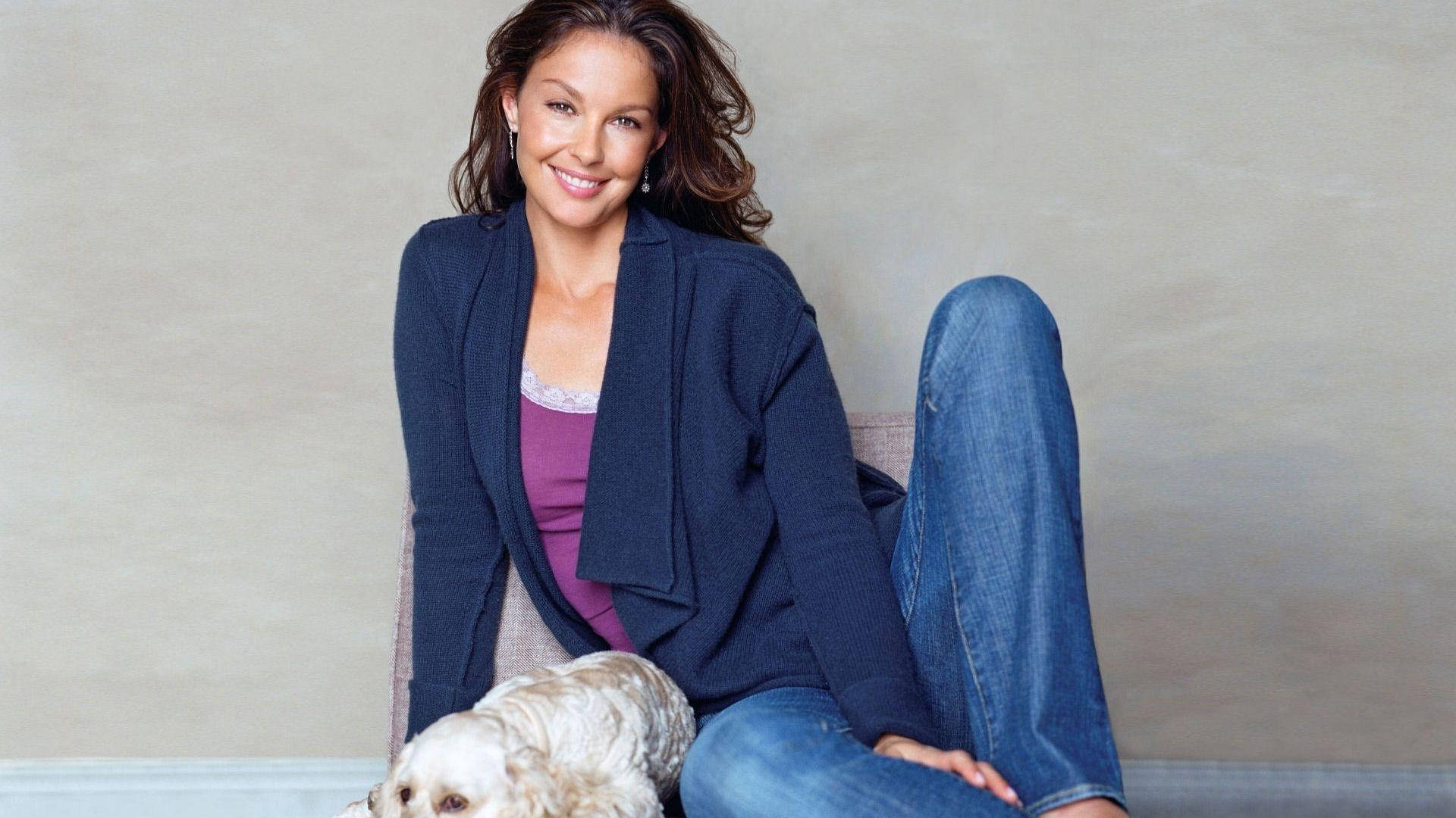 Ashley Judd 桌布 壁紙 — 一位微笑的女性穿著牛仔褲和開襟毛衣，隨意地坐在椅子上，旁邊有一隻小白狗。