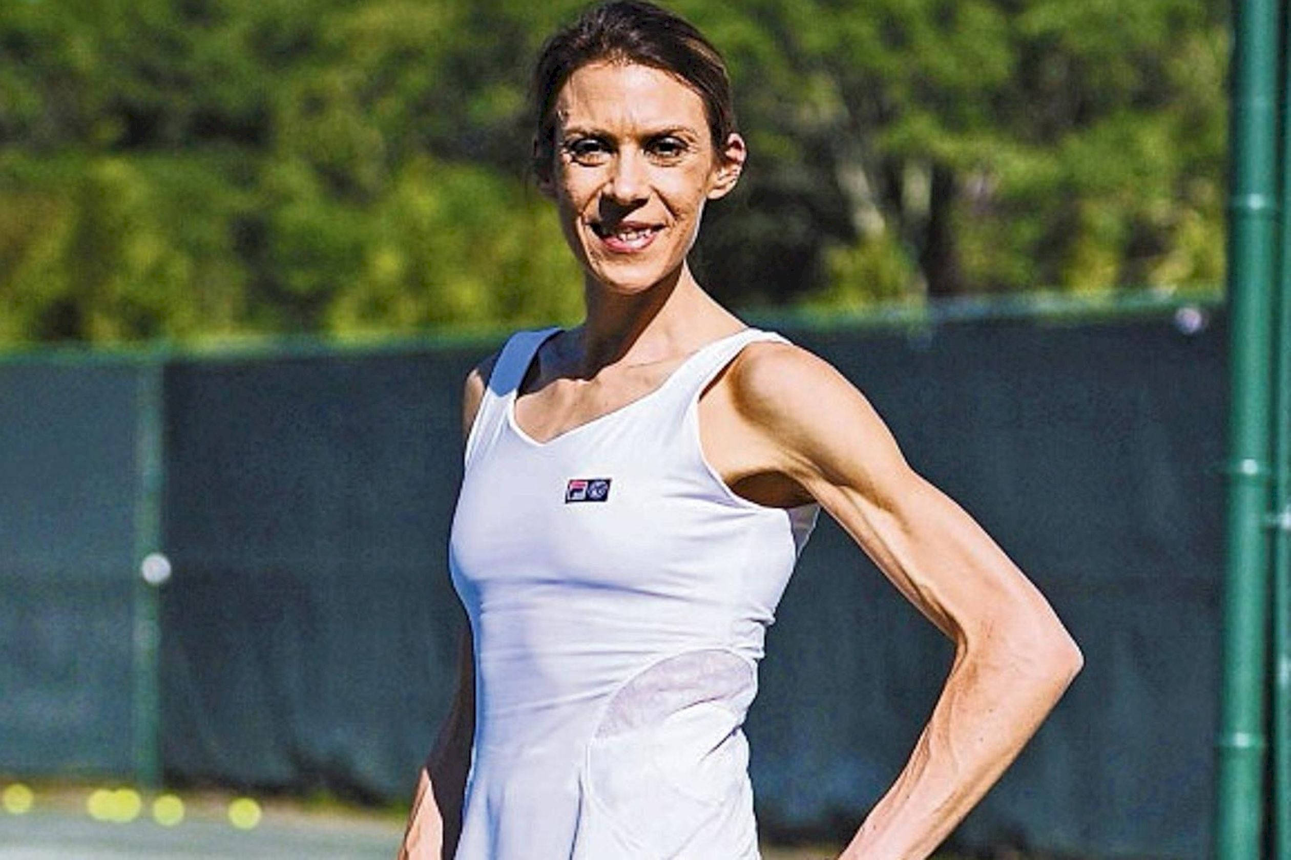 運動 桌布 壁紙 — 一位微笑的女性穿著白色網球背心，自信地站在陽光普照的球場上，身後有綠樹。