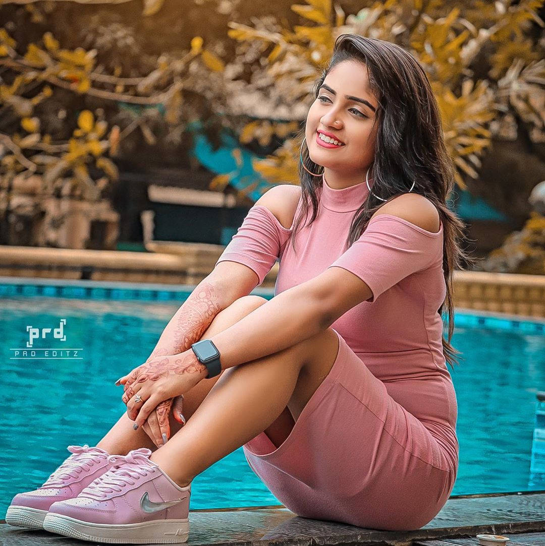 Nisha 桌布 壁紙 — 一位微笑的女性穿著粉紅色連衣裙和相配的運動鞋，坐在泳池邊，雙手有指甲花彩繪，戴著金色耳環。