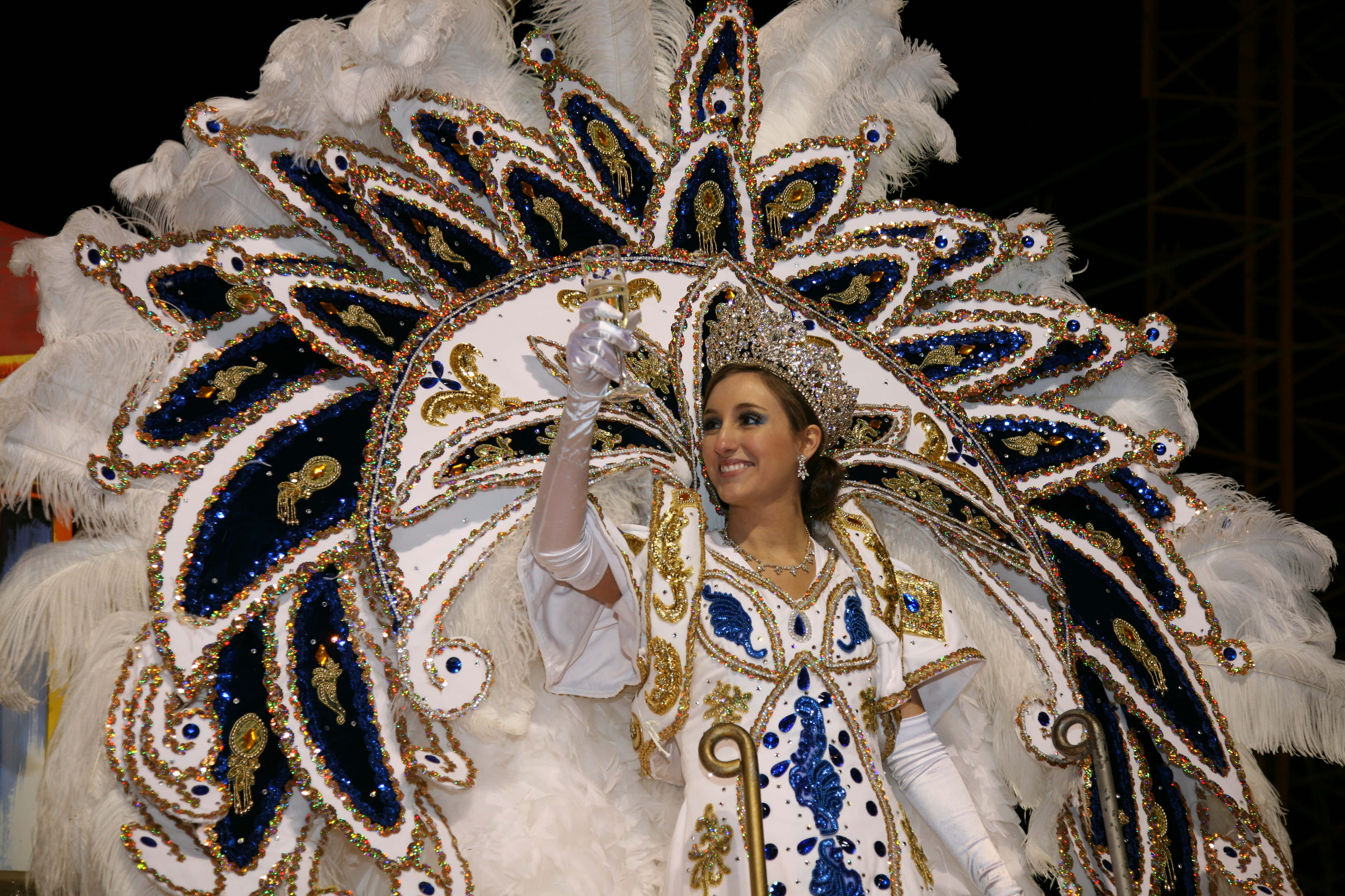 Mardi Gras 桌布 壁紙 — 一位微笑的女性穿著精緻的白色、藍色和金色羽毛嘉年華服裝，在夜晚向人群揮手。