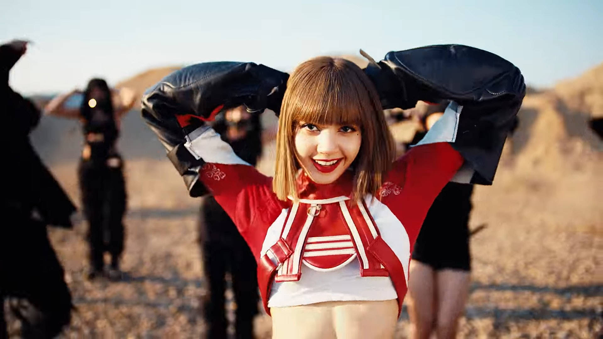 Lalisa 桌布 壁紙 — 一位微笑的女性穿著紅白賽車服，在陽光明媚的沙漠背景下俏皮地舉起一件黑色皮夾克。