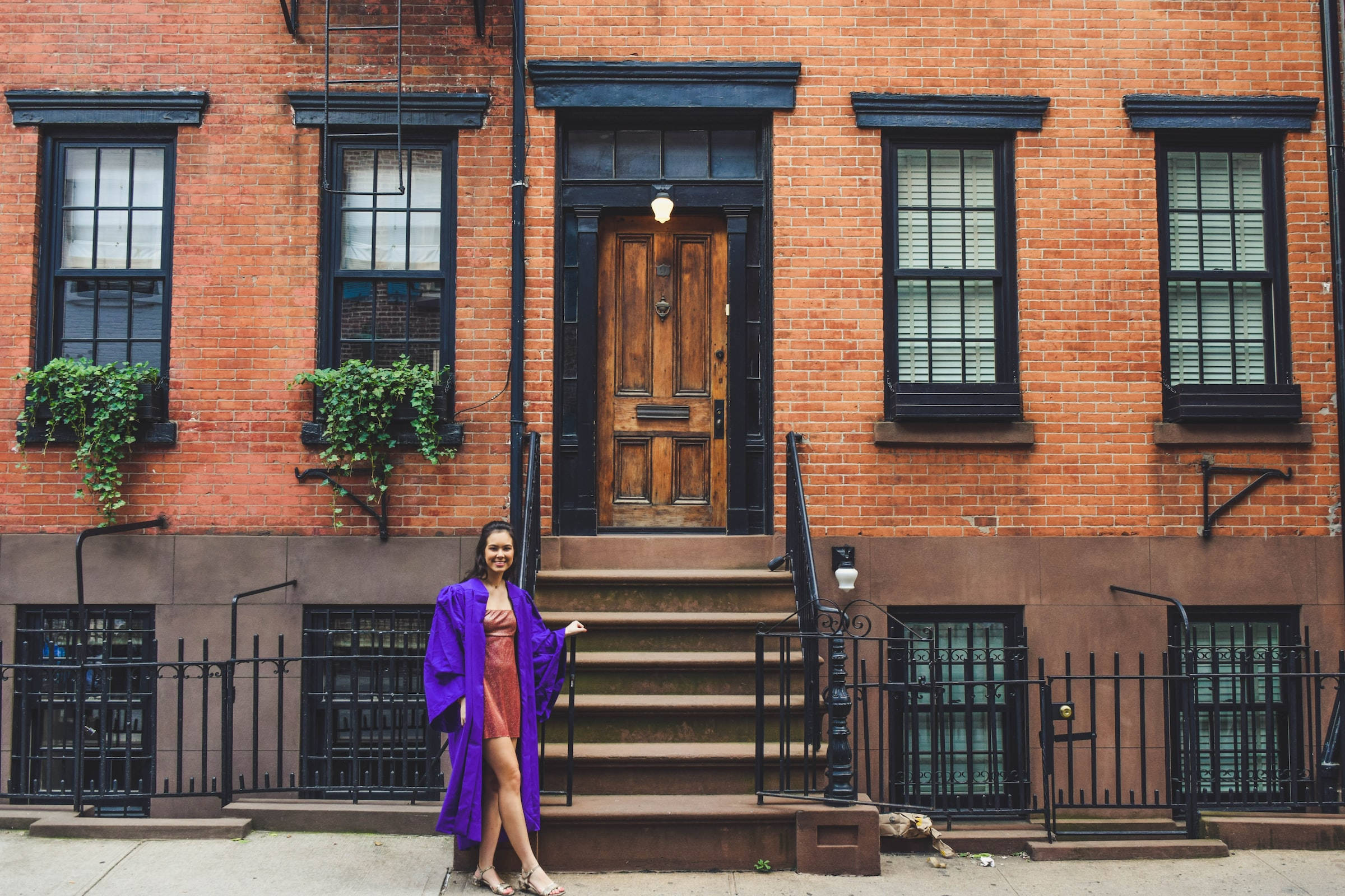 Graduation 桌布 壁紙 — 一位微笑的女性穿著紫色畢業袍，站在經典紅磚連棟房屋的階梯上，階梯有黑色飾邊和常春藤。